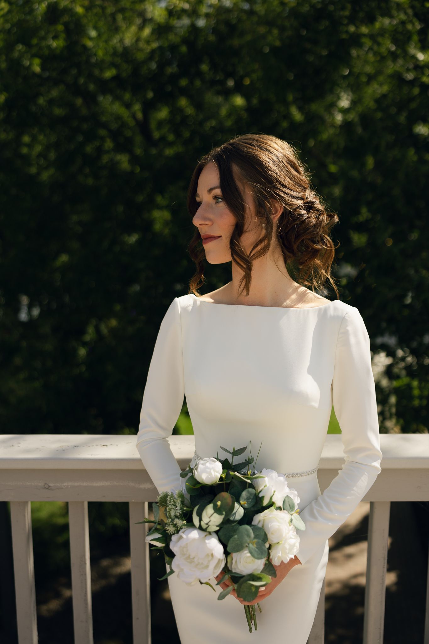 Bride in classic white long-sleeve dress holds bouquet of white roses on sunny balcony.