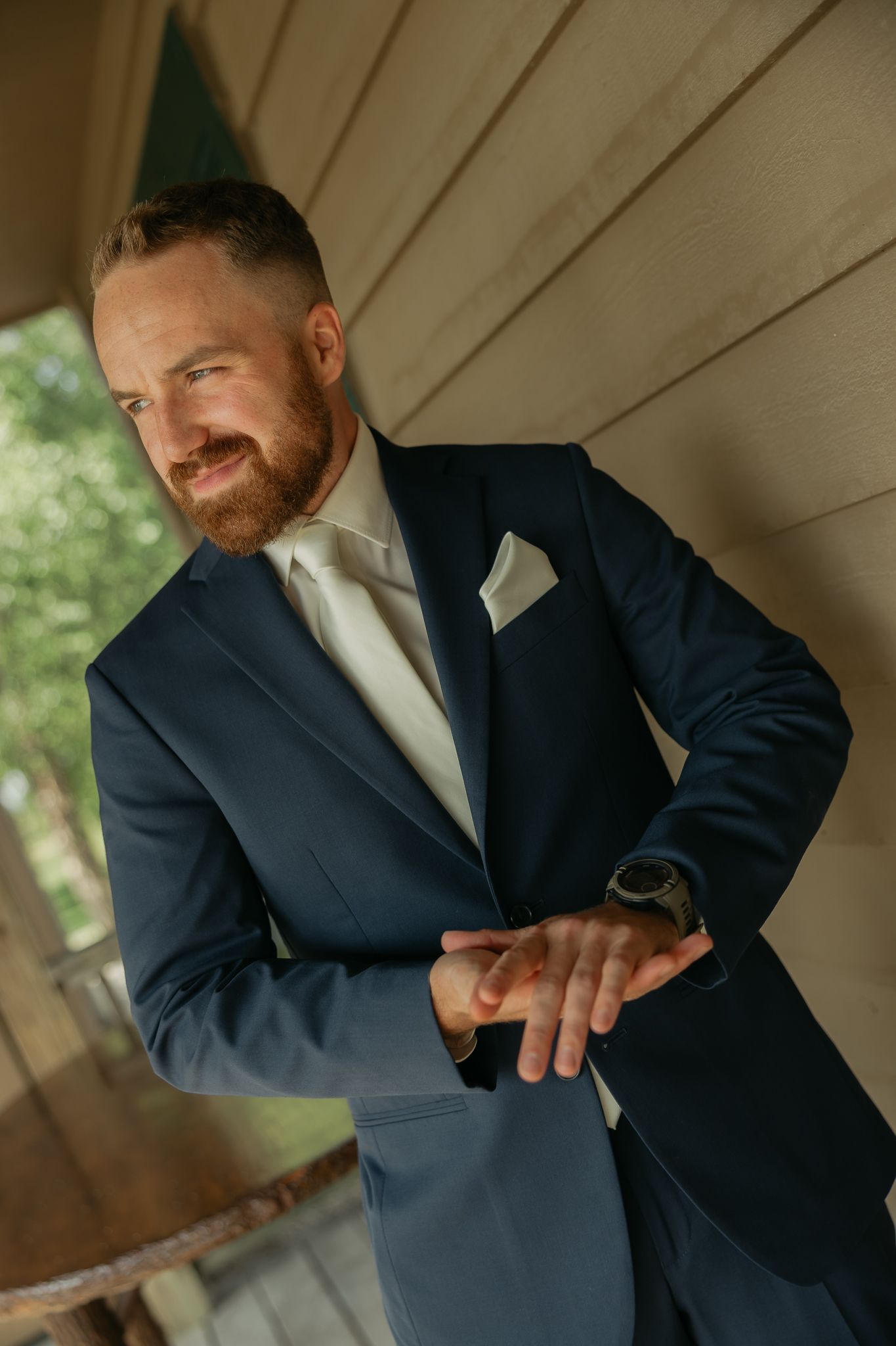 A groom in a navy suit adjusts his tie while standing on a covered porch.