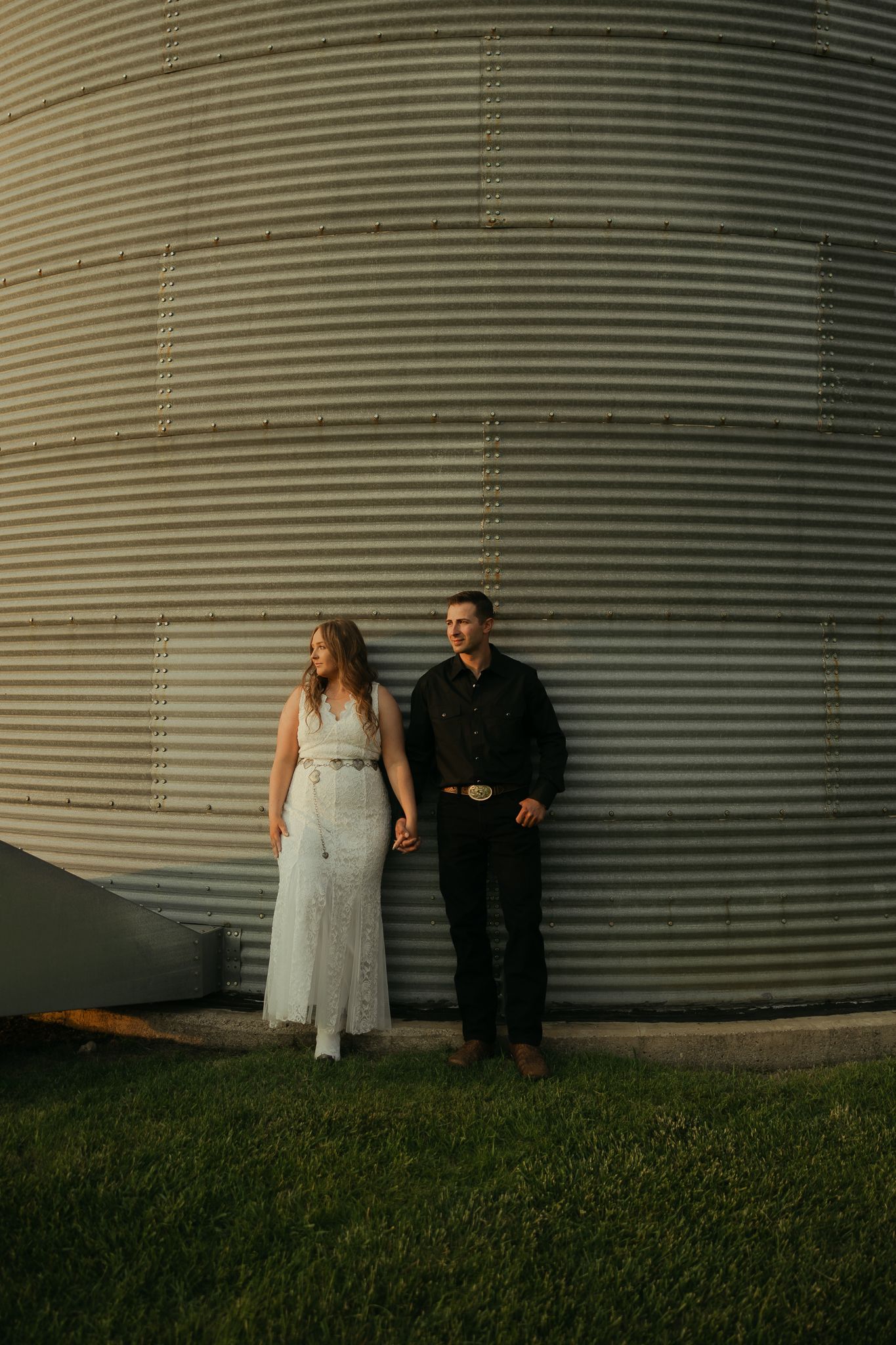 Wedding couple poses against curved metal grain silo in dramatic lighting.