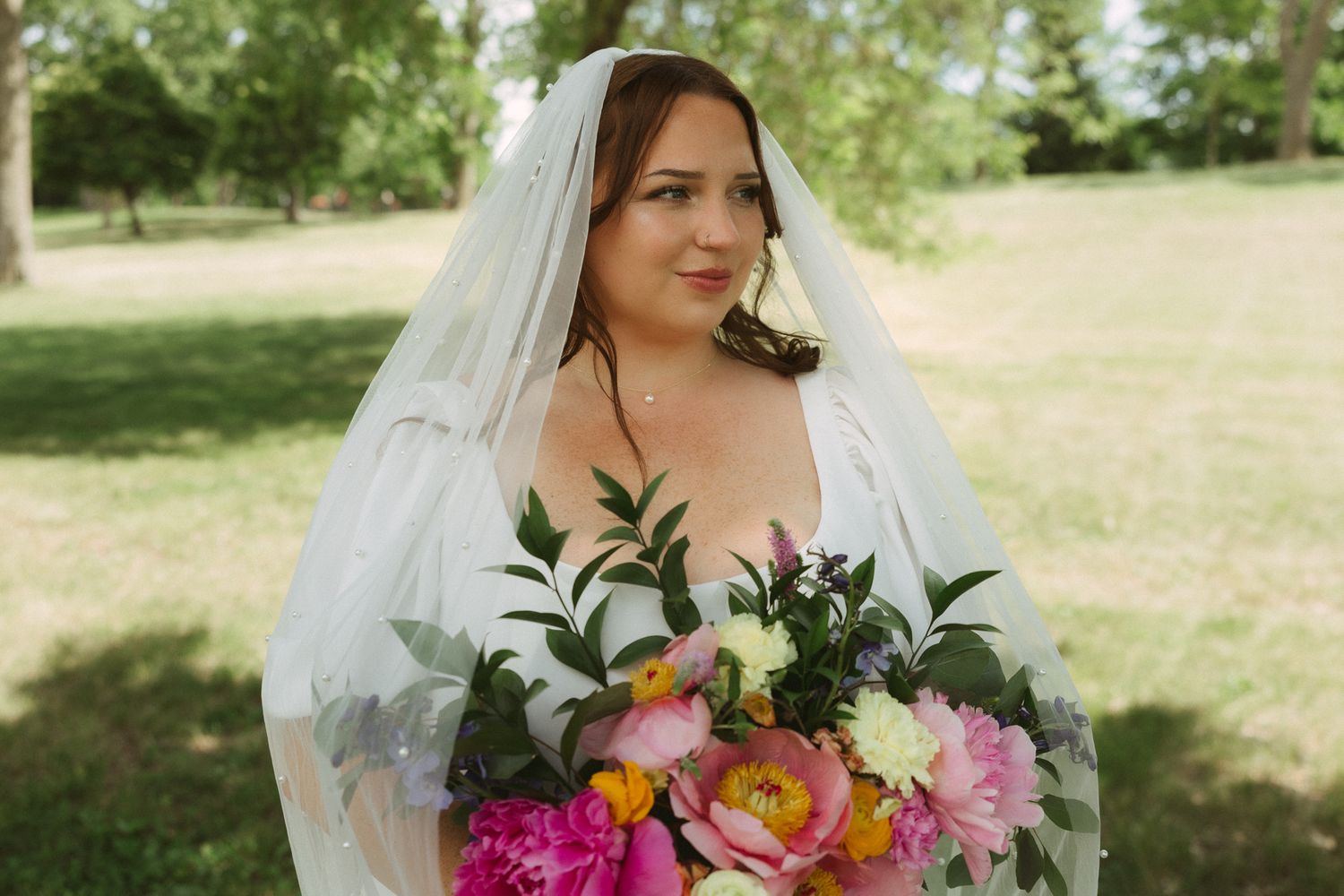 Bride in white veil holding vibrant pink and yellow floral bouquet outdoors in lush green park setting.