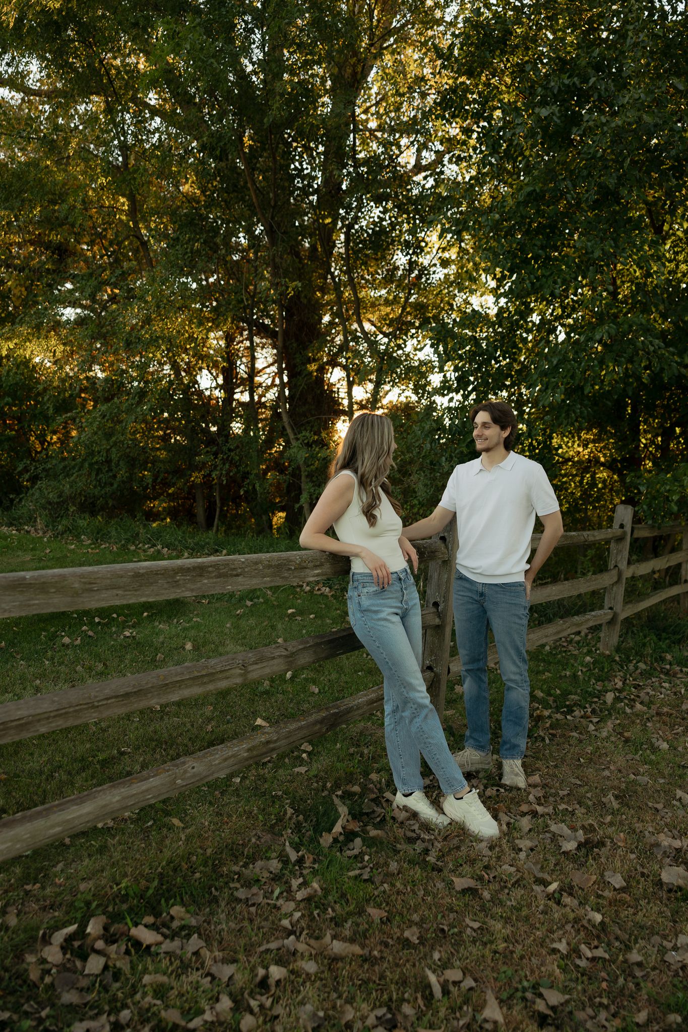 Couple in casual wear leaning against a wooden fence at sunset surrounded by trees.