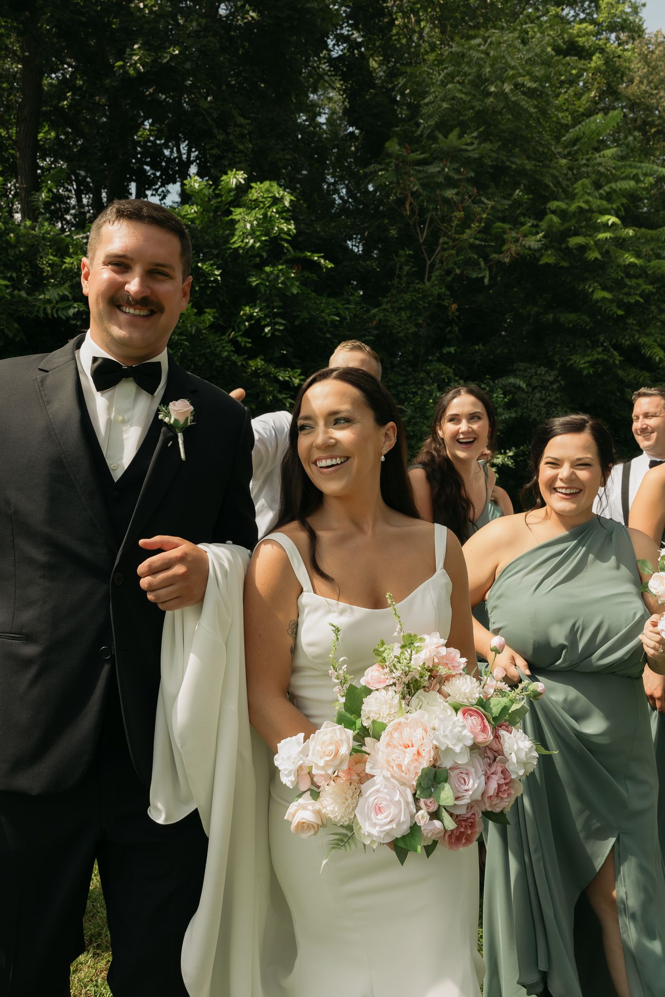 Wedding party members smiling outdoors with pink floral bouquet.