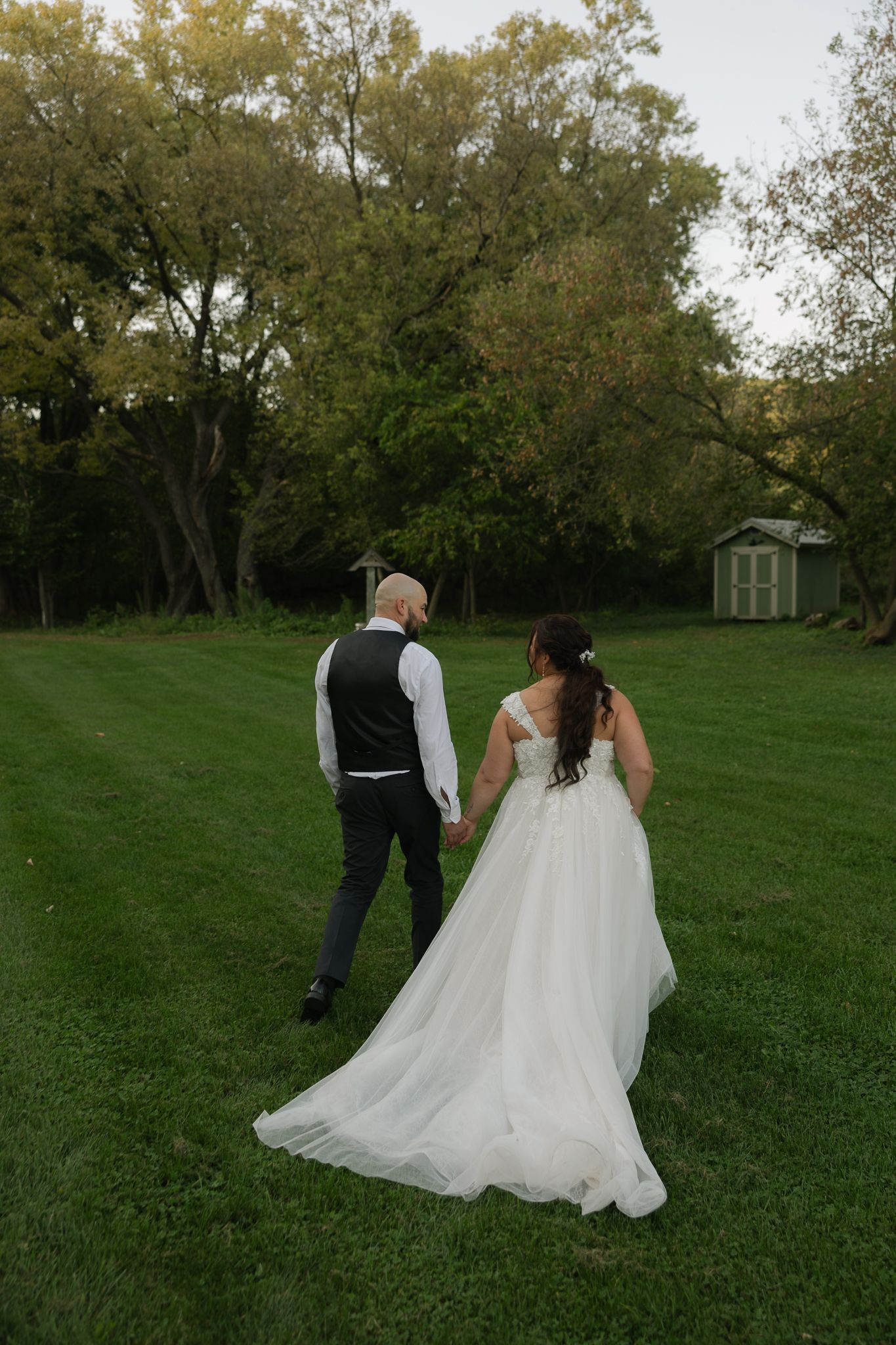 Couple walking through a green field with white dress flowing behind them.