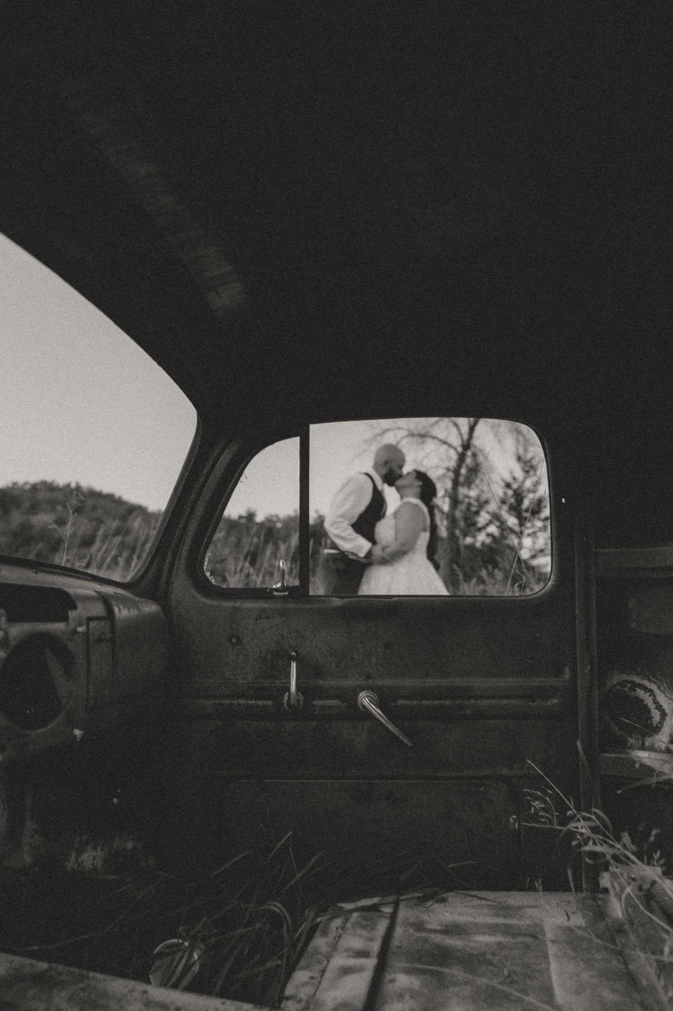 Black and white view through vintage car window of wedding couple in field.