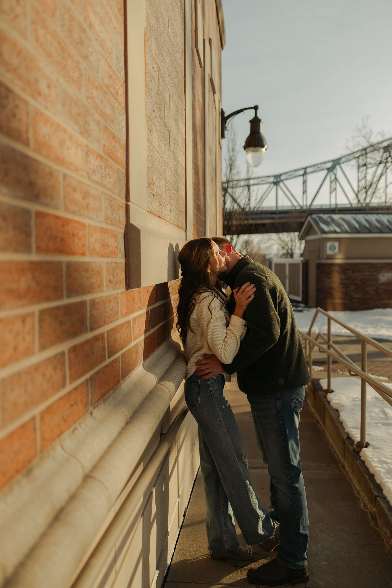 Romantic couple embracing against a brick wall with industrial bridge in background at sunset.