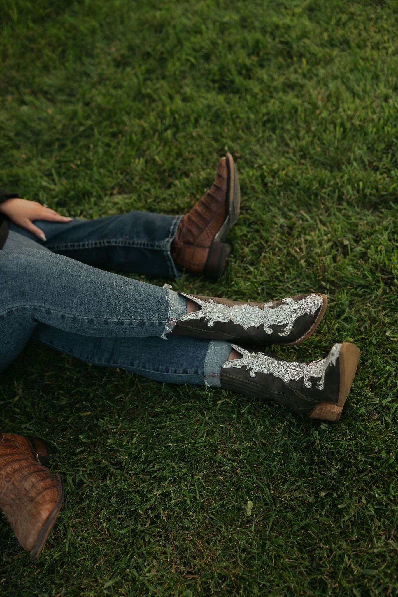 Distressed denim jeans and brown leather boots rest on green grass during an outdoor photoshoot.
