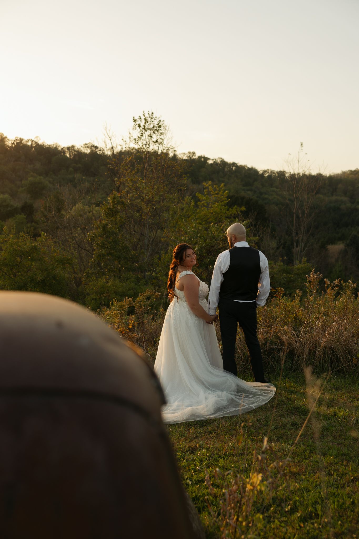 Newlyweds share an intimate moment at sunset in a rustic outdoor setting with flowing white dress and autumn foliage.