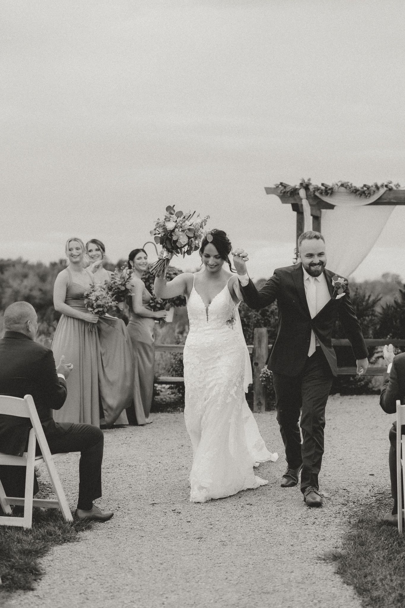 Black and white photo of wedding party celebrating on gravel path after ceremony.