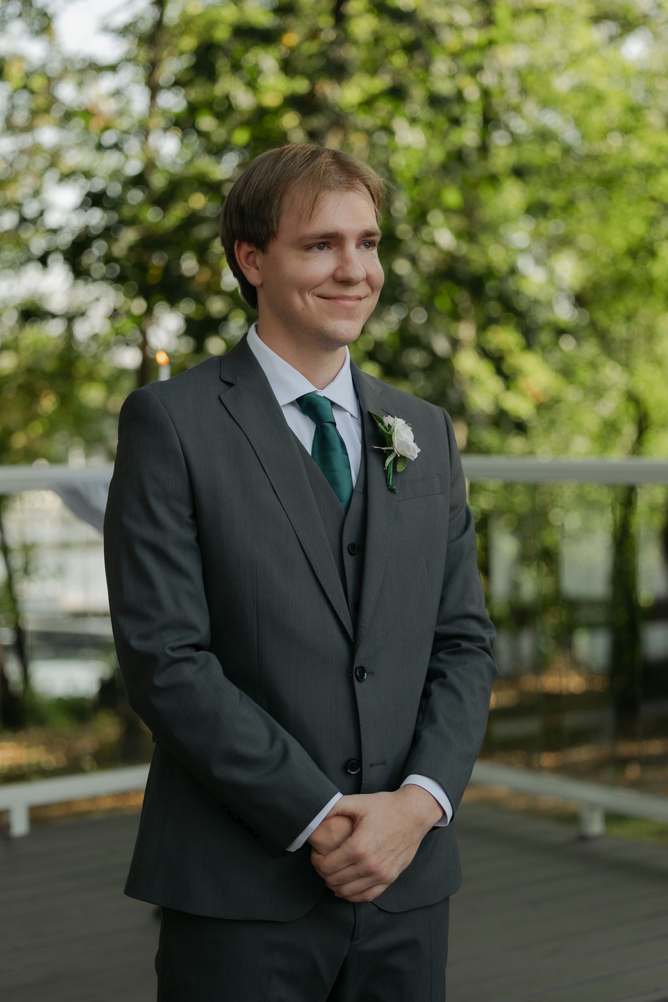 Groom in dark suit standing outdoors with trees in background.