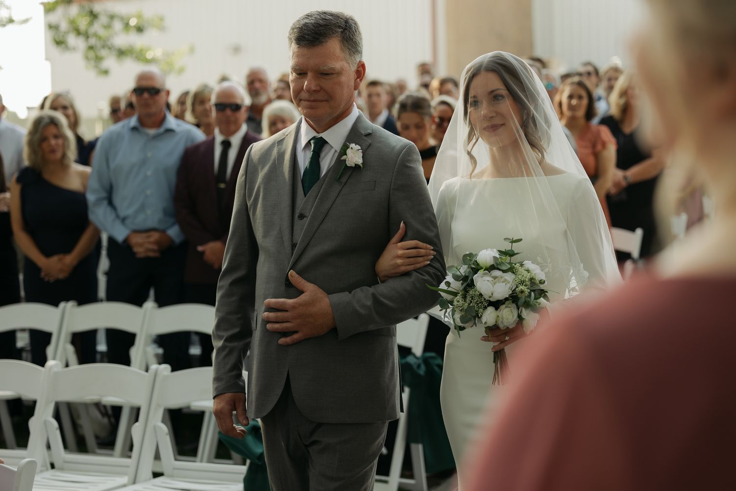 Indoor wedding ceremony with guests seated in white chairs and natural lighting.