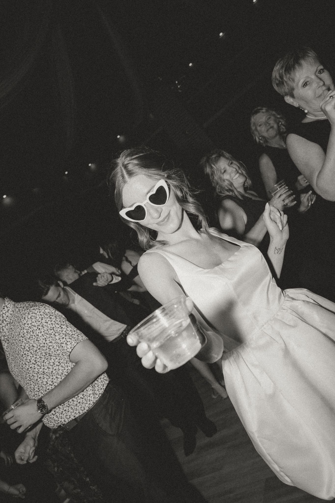Black and white photo of guest in white dress and sunglasses holding drink on dance floor.