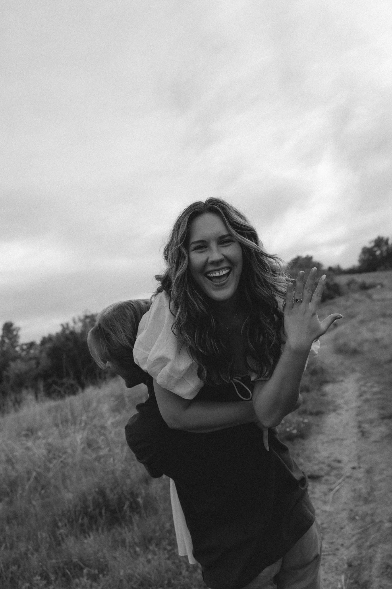 Black and white photo of a laughing woman carrying a child on her back outdoors on a rural path.