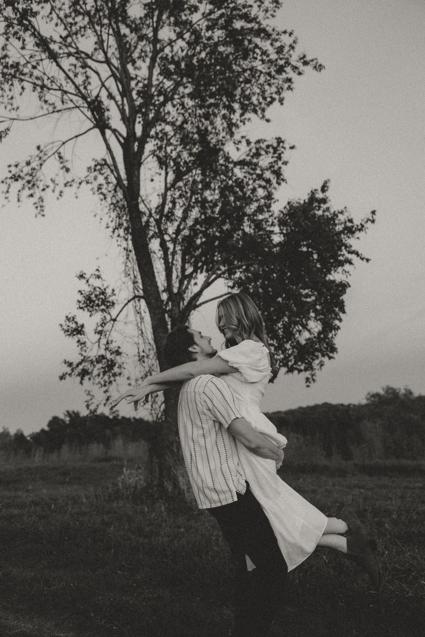 Playful couple embracing and spinning in a field near a lone tree.