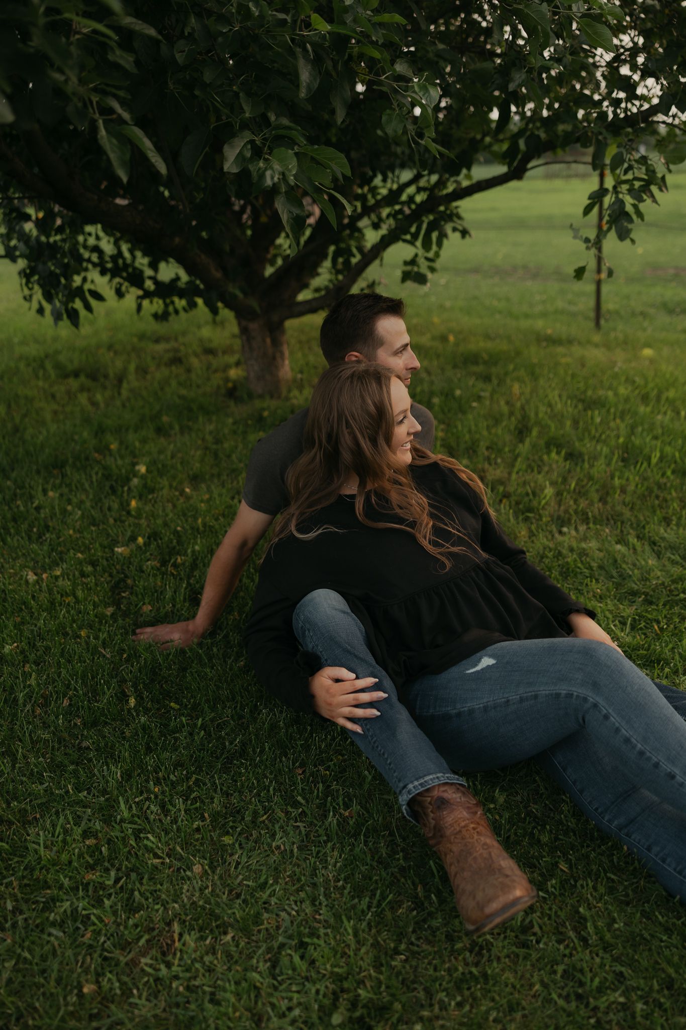 A couple relaxes on lush grass beneath a leafy tree in a romantic outdoor setting.