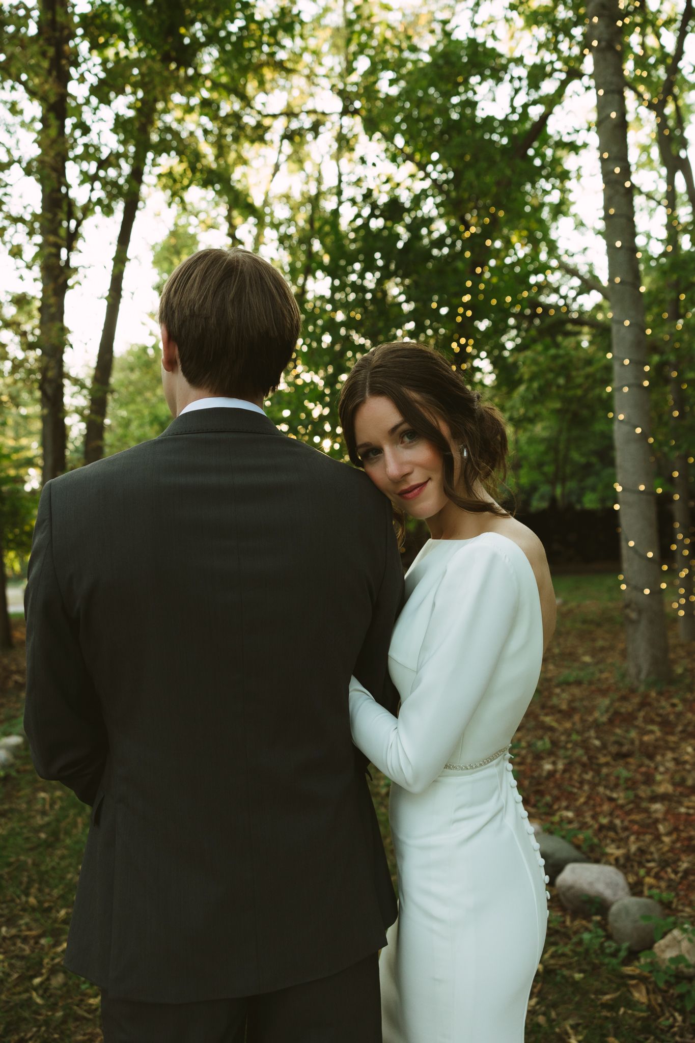 A couple in wedding attire embraces while standing among autumn trees with fallen leaves in a romantic outdoor setting.