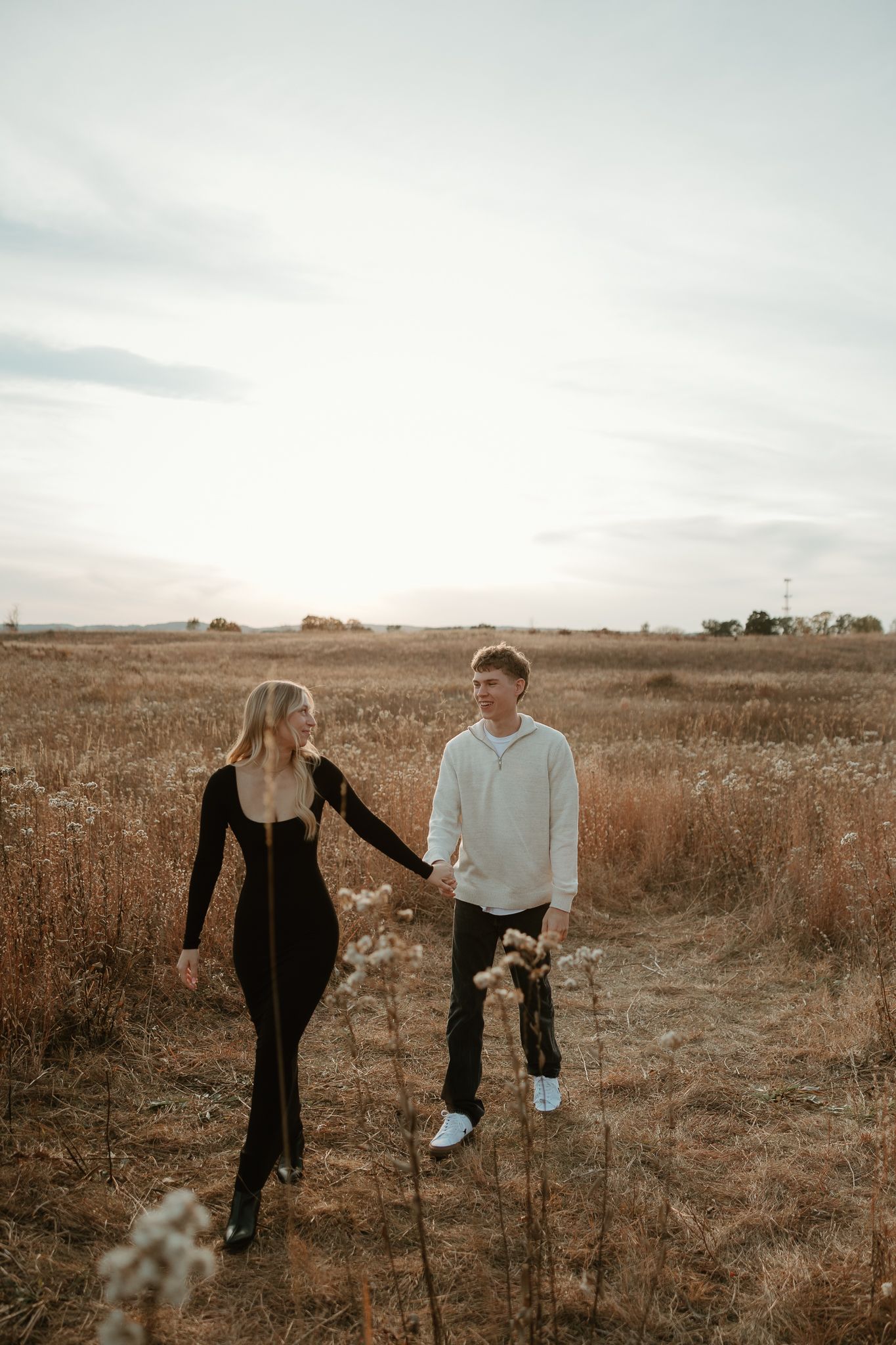 Couple walks through autumn field at sunset carrying coffee cups during engagement shoot.