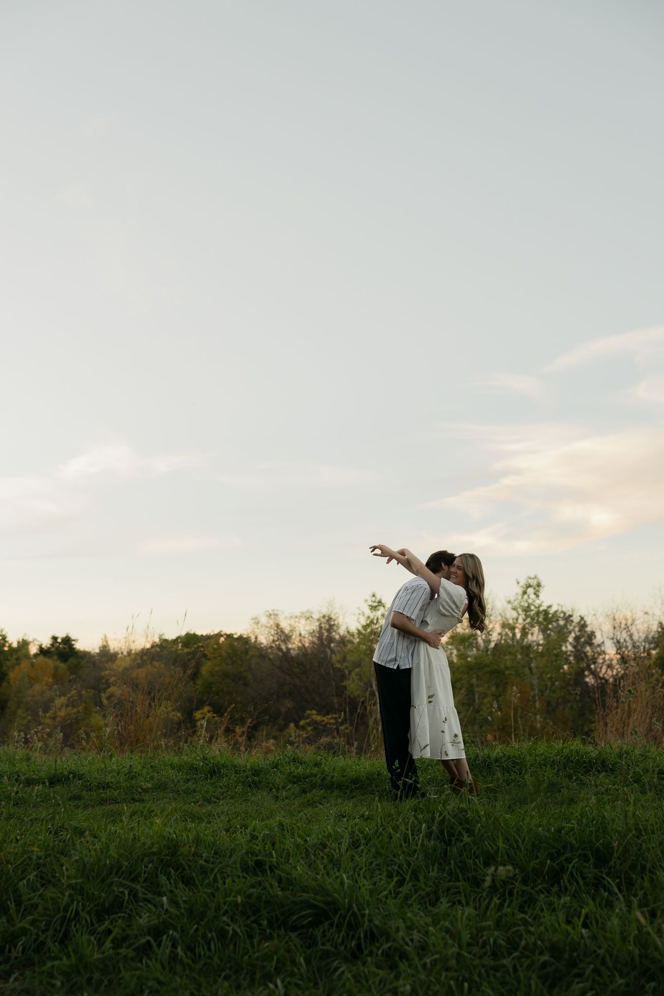A romantic couple embraces in a grassy field at sunset with trees in the background.