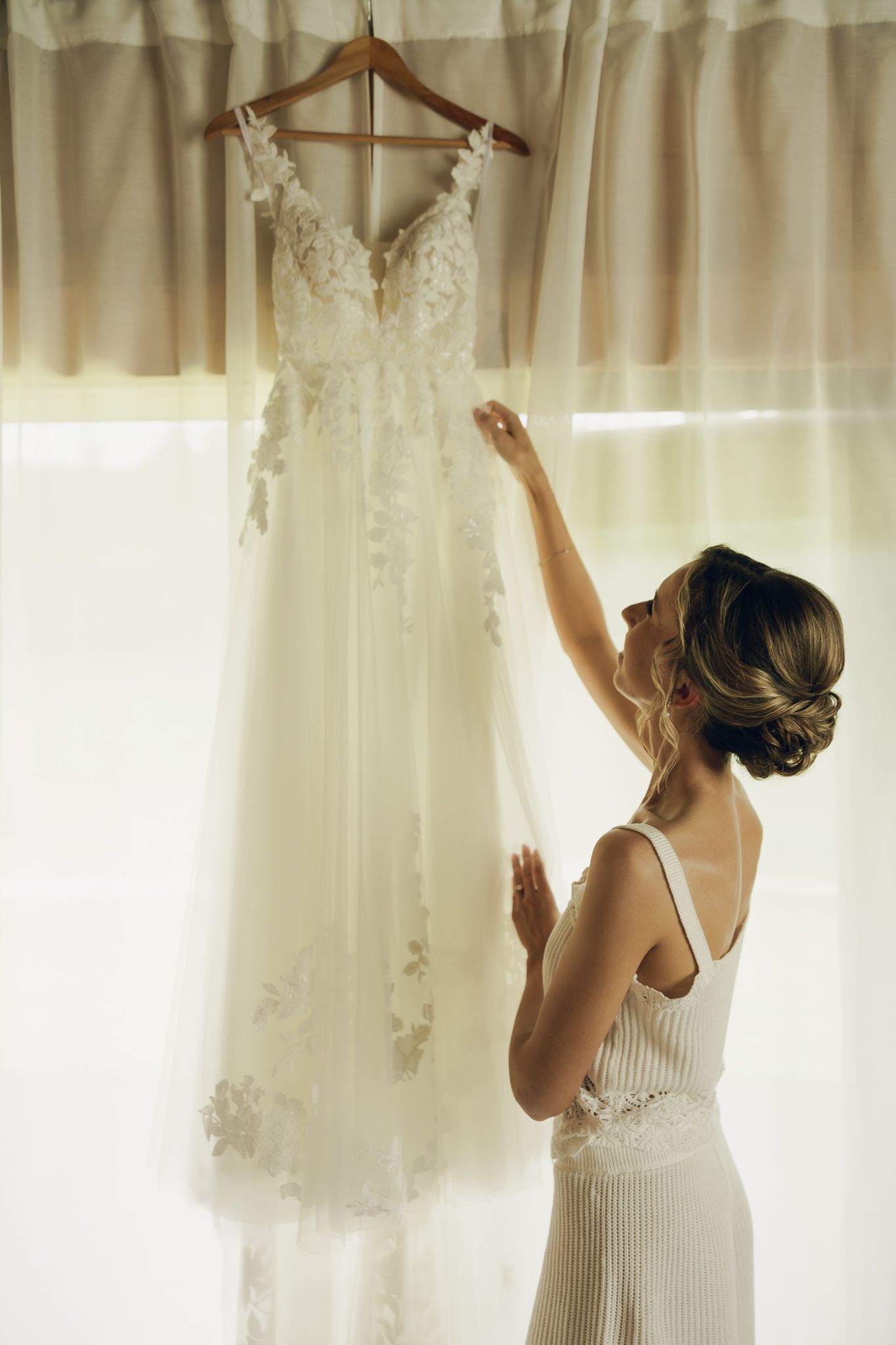 Person in white dress reaching up to adjust wedding gown hanging on display.