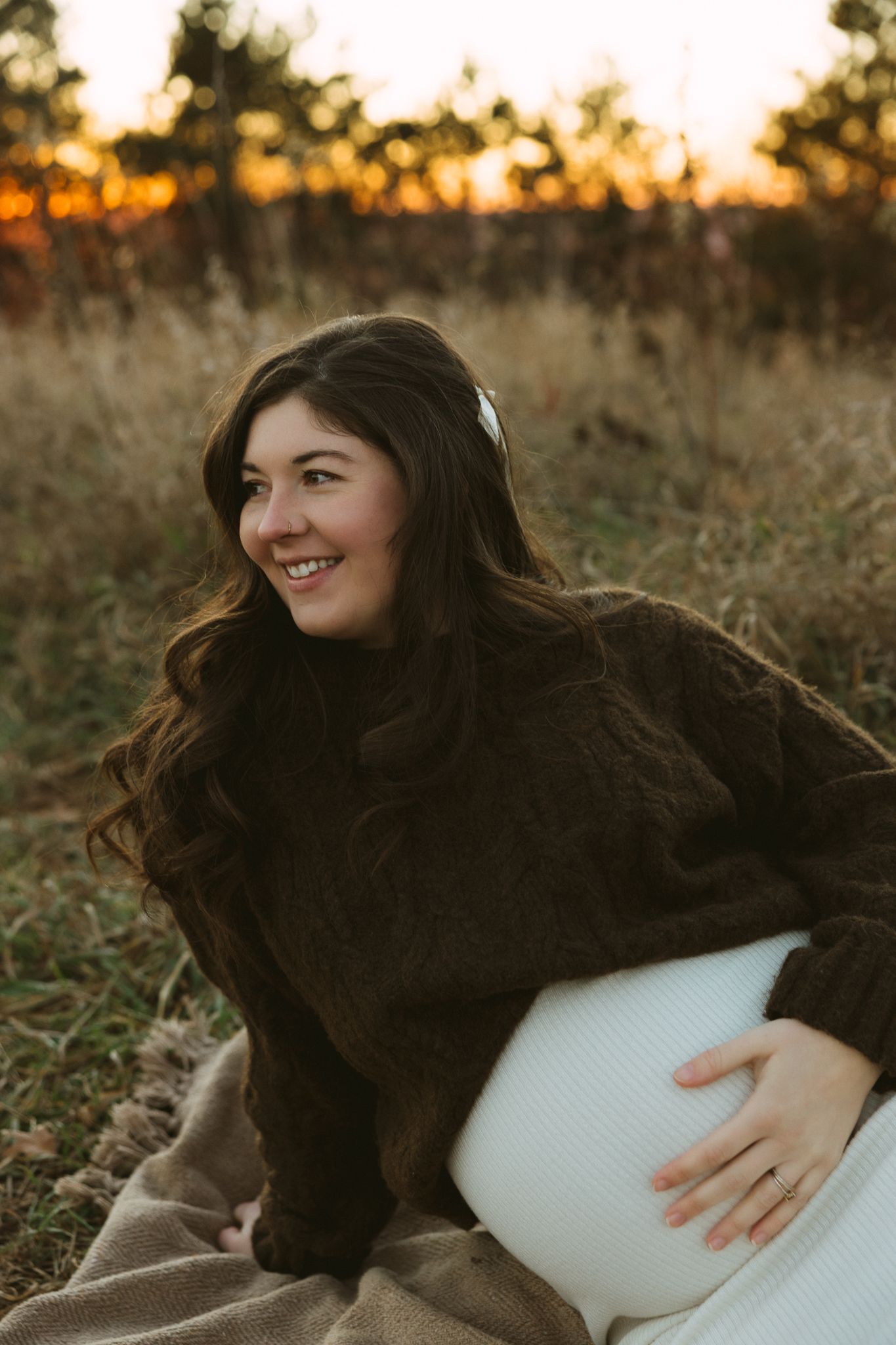A person in a brown sweater sits on a blanket holding a white pillow at sunset in a grassy field.
