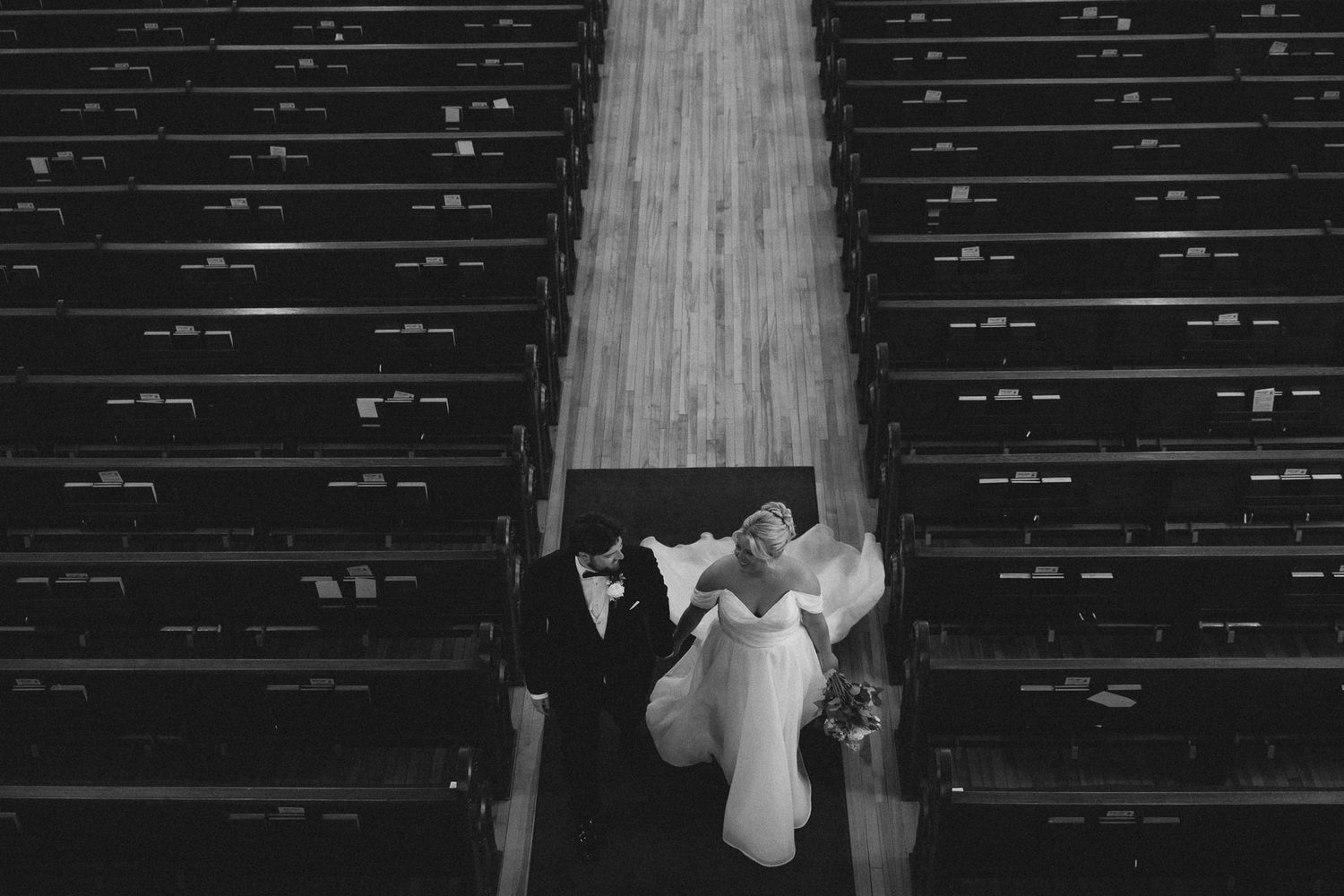 Black and white photo of bride and groom walking down church aisle surrounded by empty wooden pews.