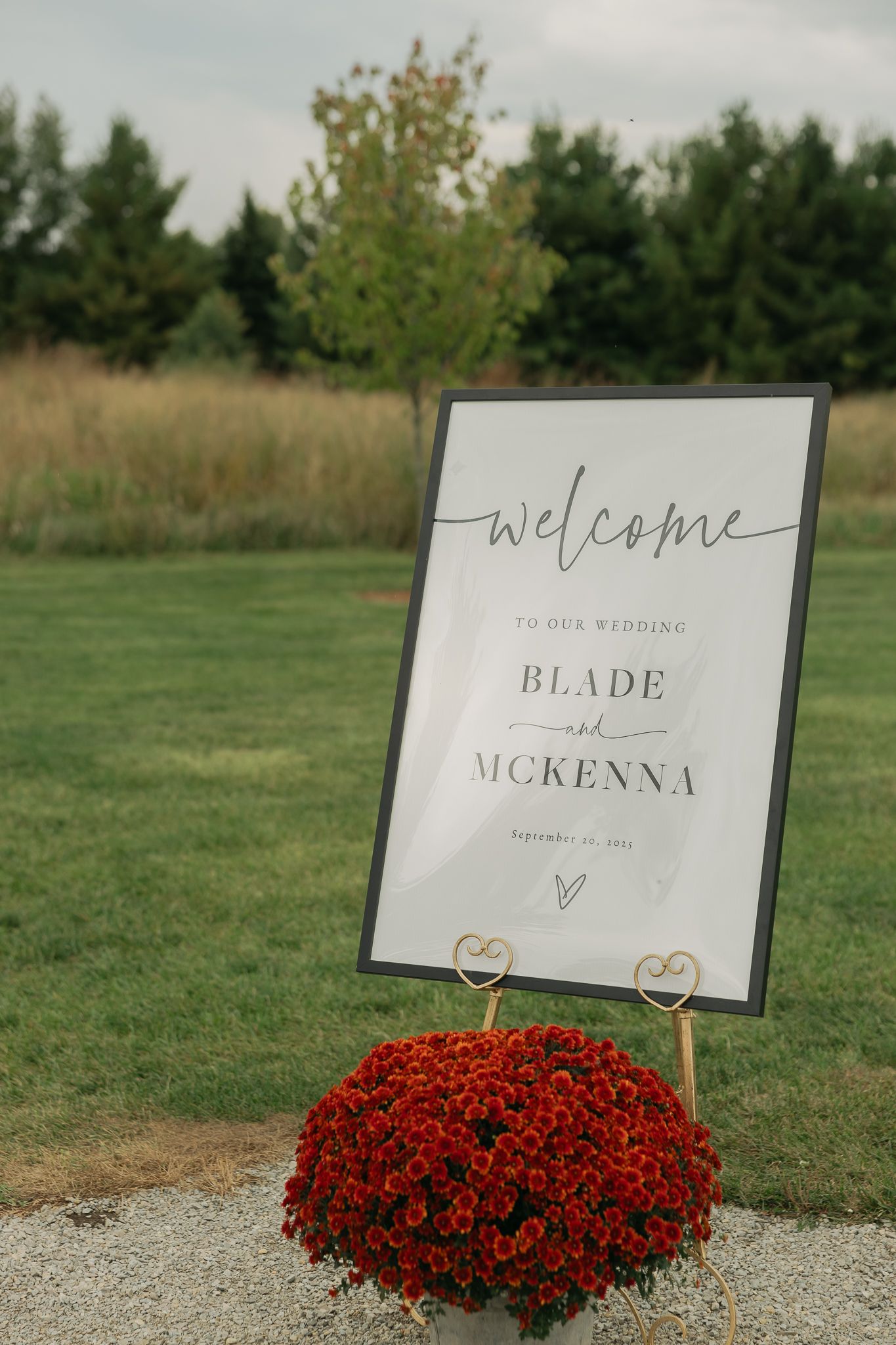 Wedding welcome signs and red mum flowers line a gravel path at an outdoor venue.