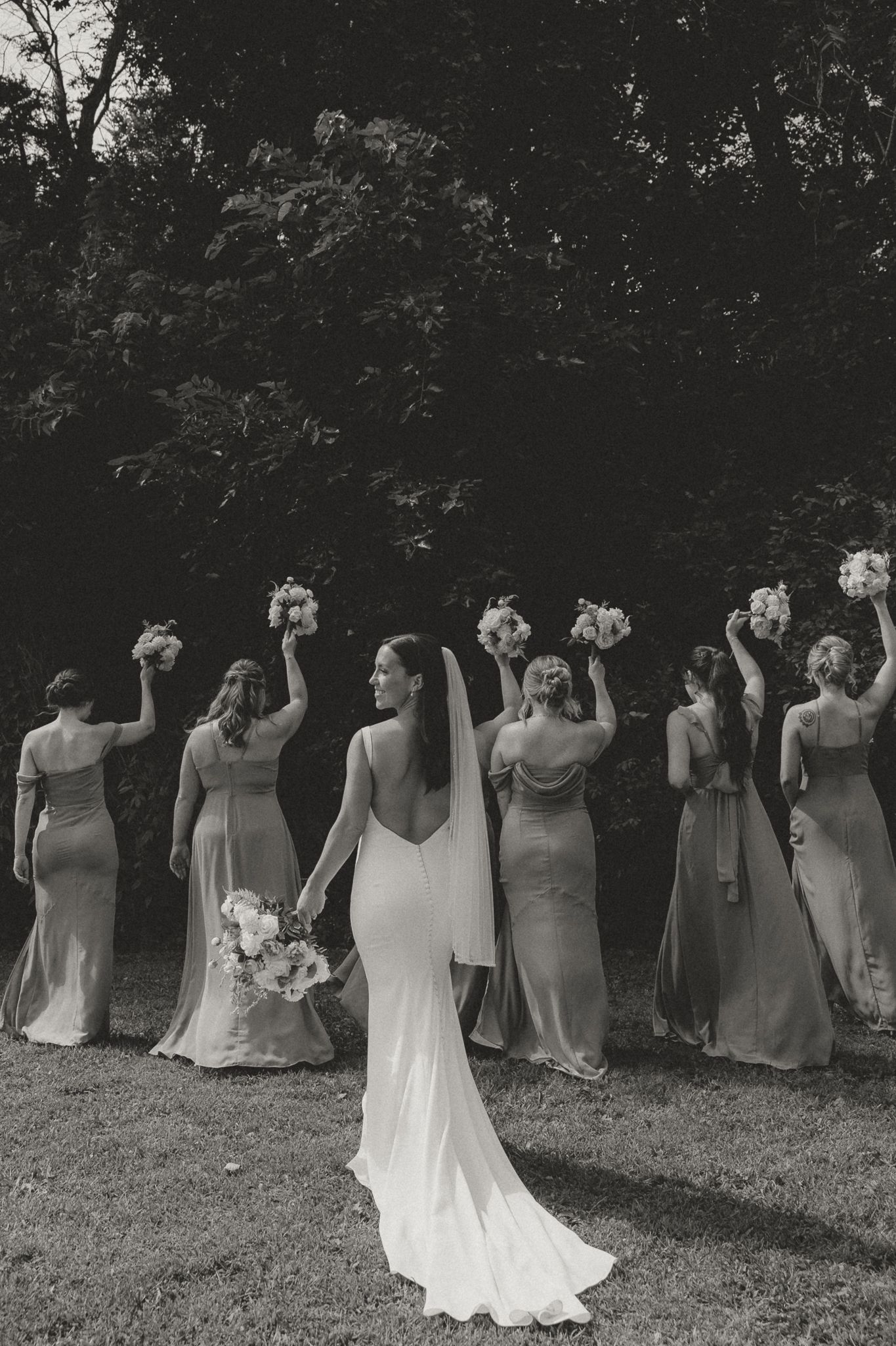 Artistic black and white photo of bridesmaids in elegant dresses holding bouquets against forest backdrop.