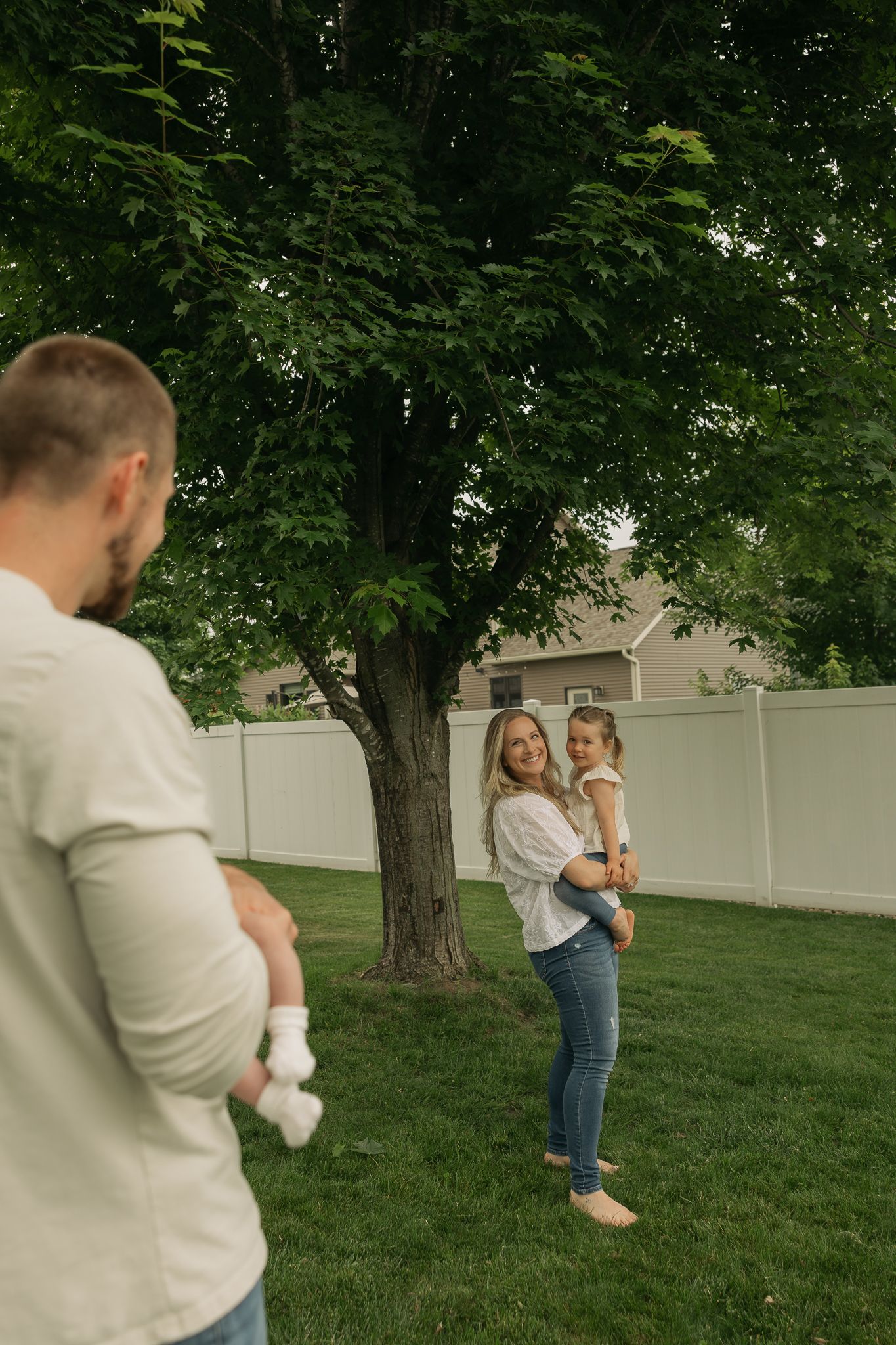 Family playing together in a backyard with white fence and large trees.