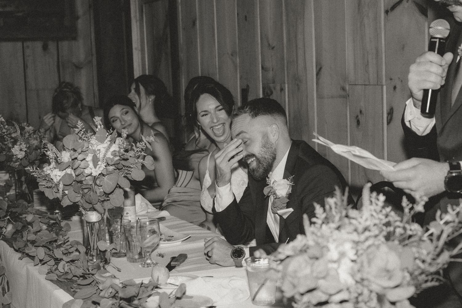 Black and white reception photo showing wedding guests laughing at a decorated dinner table.