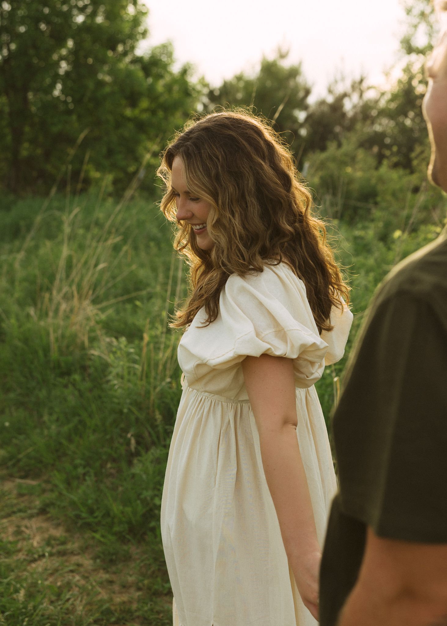 A woman in a white dress smiles softly in a sunlit green field, her long brown hair glowing in golden hour light.