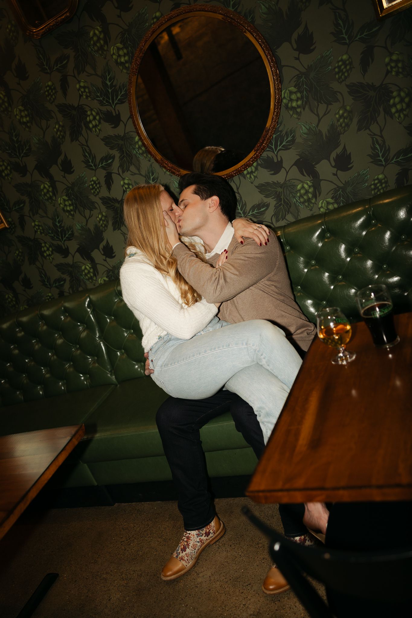 Couple embracing on a green leather booth with decorative wallpaper and string lights in background.