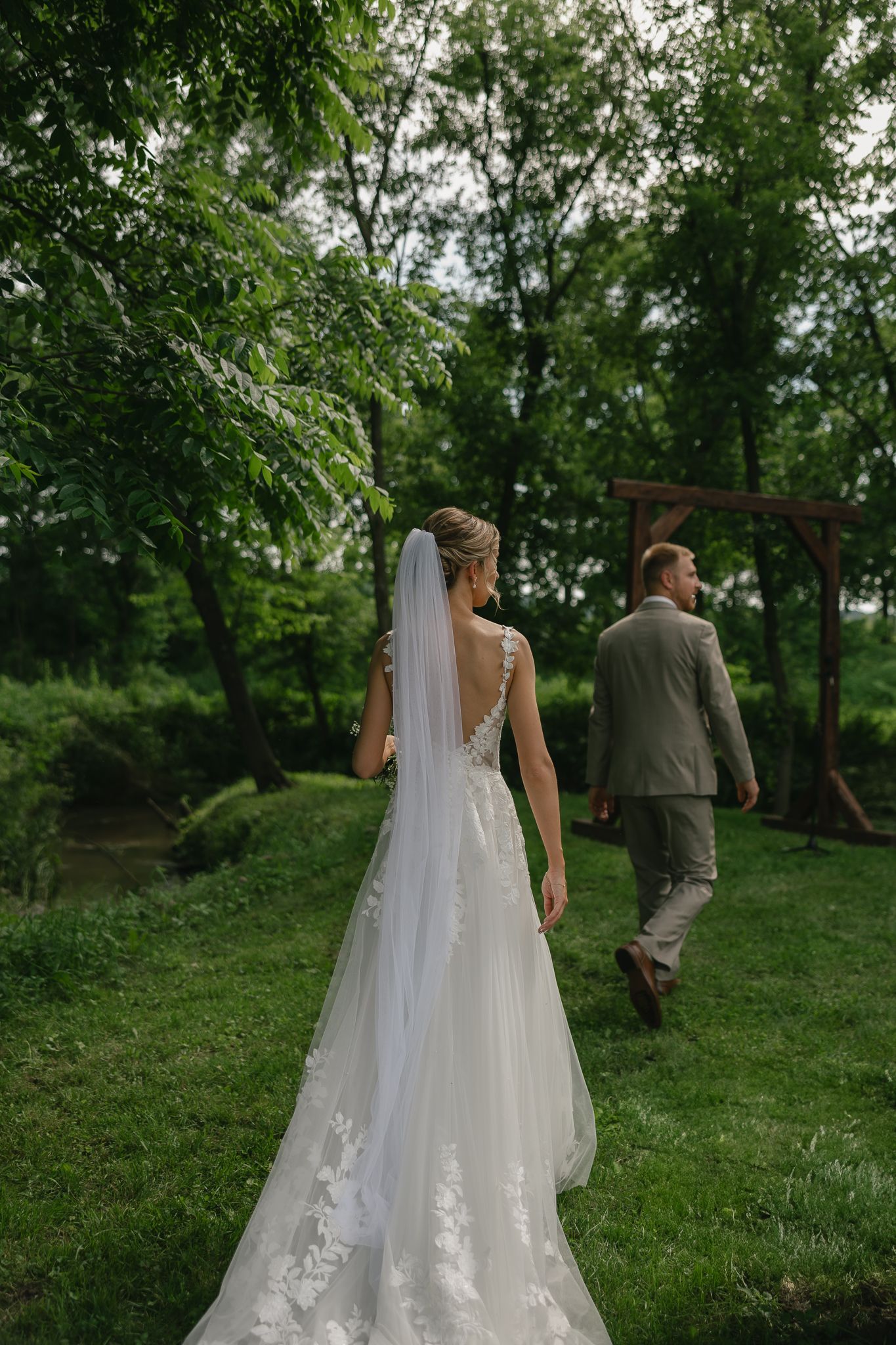 A couple walks through a lush green garden path surrounded by trees on their wedding day.