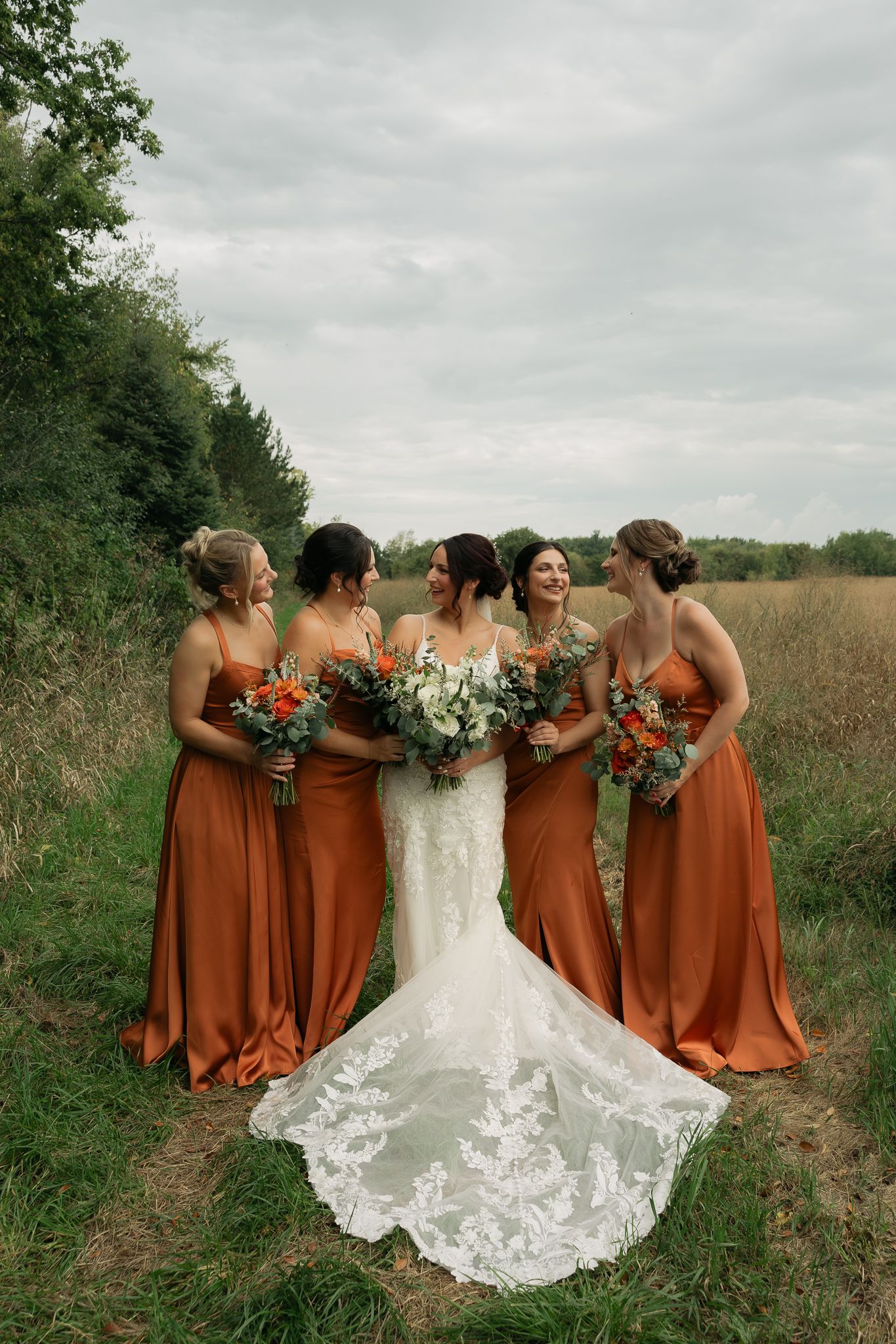 Bride in white lace gown stands with bridesmaids wearing rust-colored dresses in a field.
