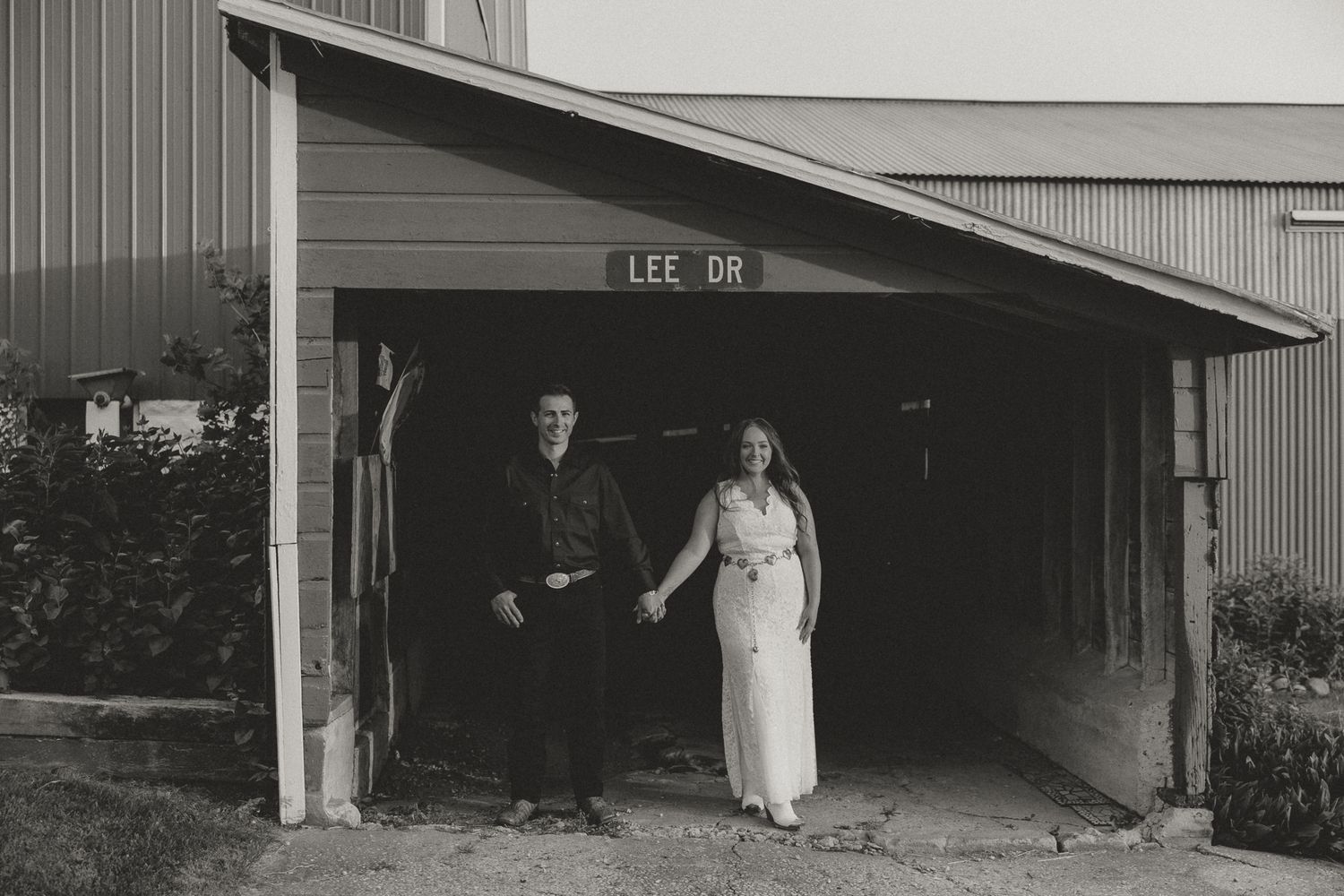 A couple stands under a rustic barn overhang in formal wear for a black and white photo.