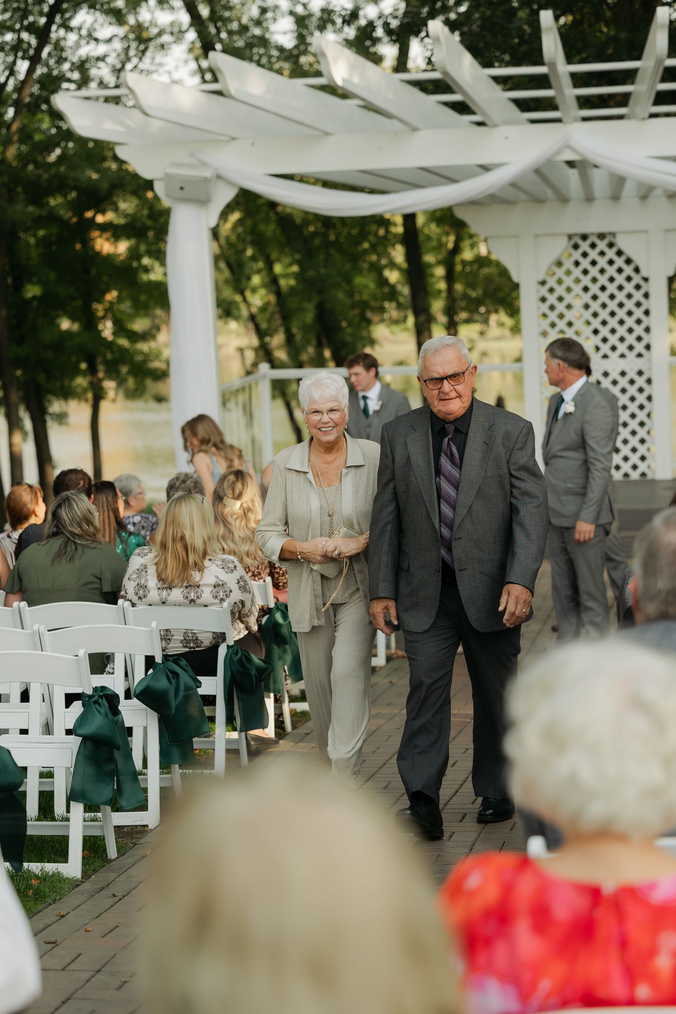 Guests arrive at an outdoor wedding venue decorated with white chairs and red flowers.