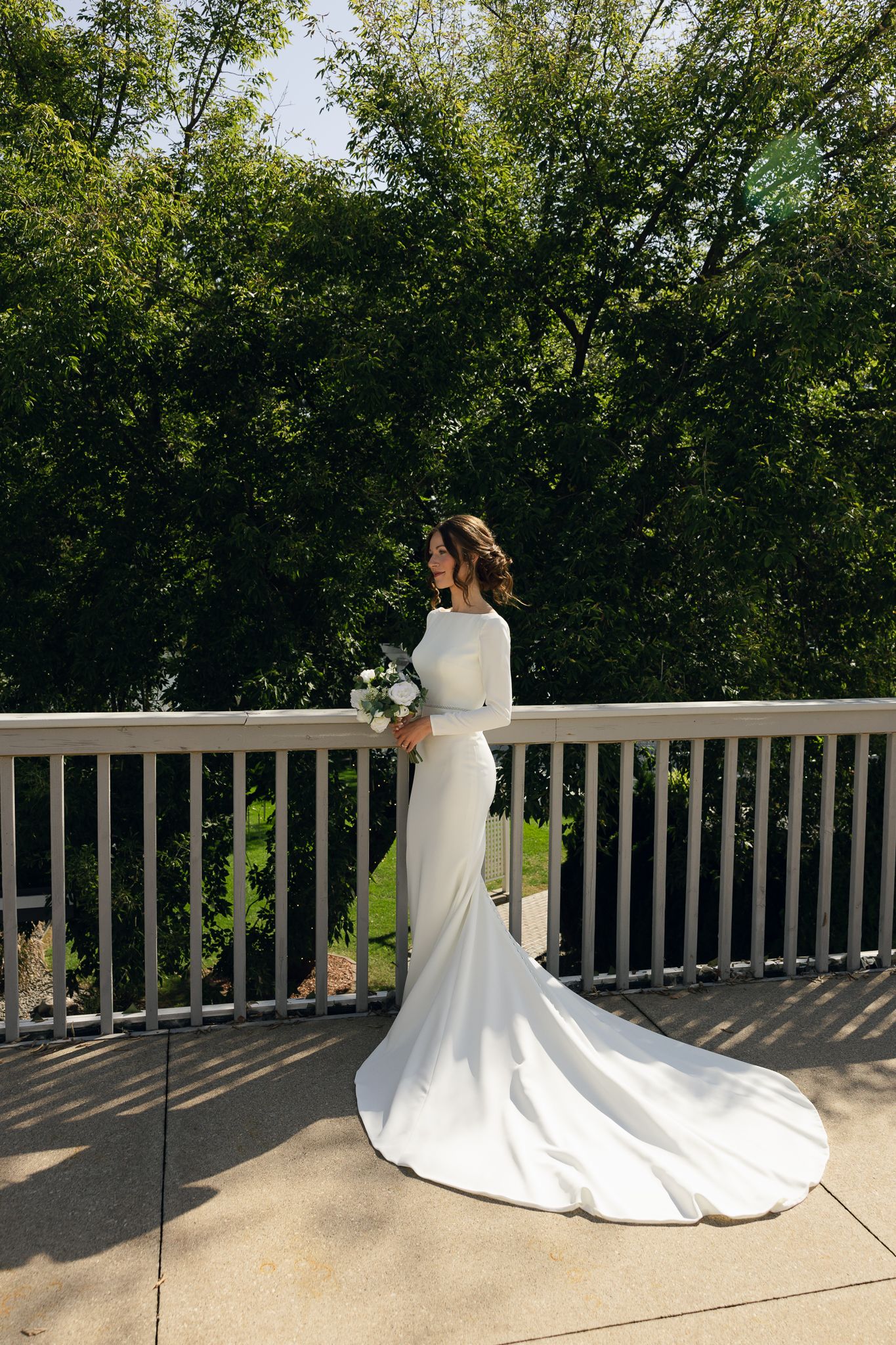 Elegant bride in fitted white long-sleeve gown with train poses on outdoor deck surrounded by trees.