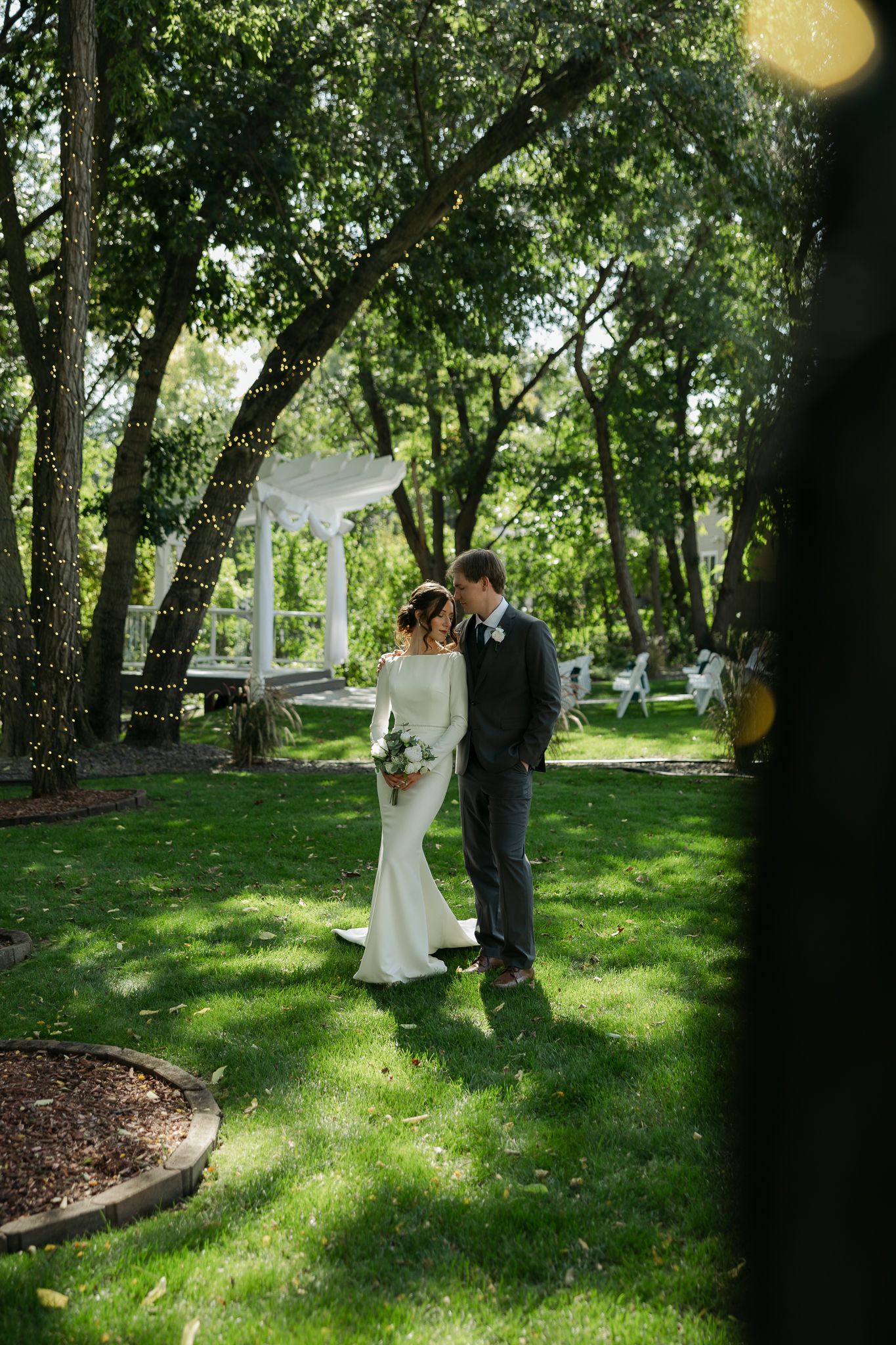 Newlyweds share a romantic moment in a sunlit garden with trees.