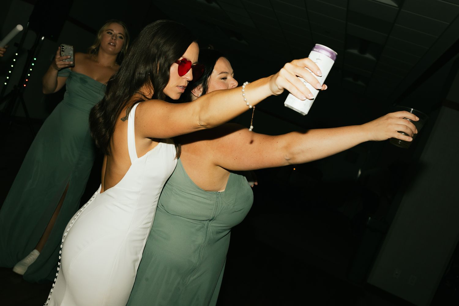 Wedding guests dance energetically at a reception while holding drinks in a dimly lit venue.