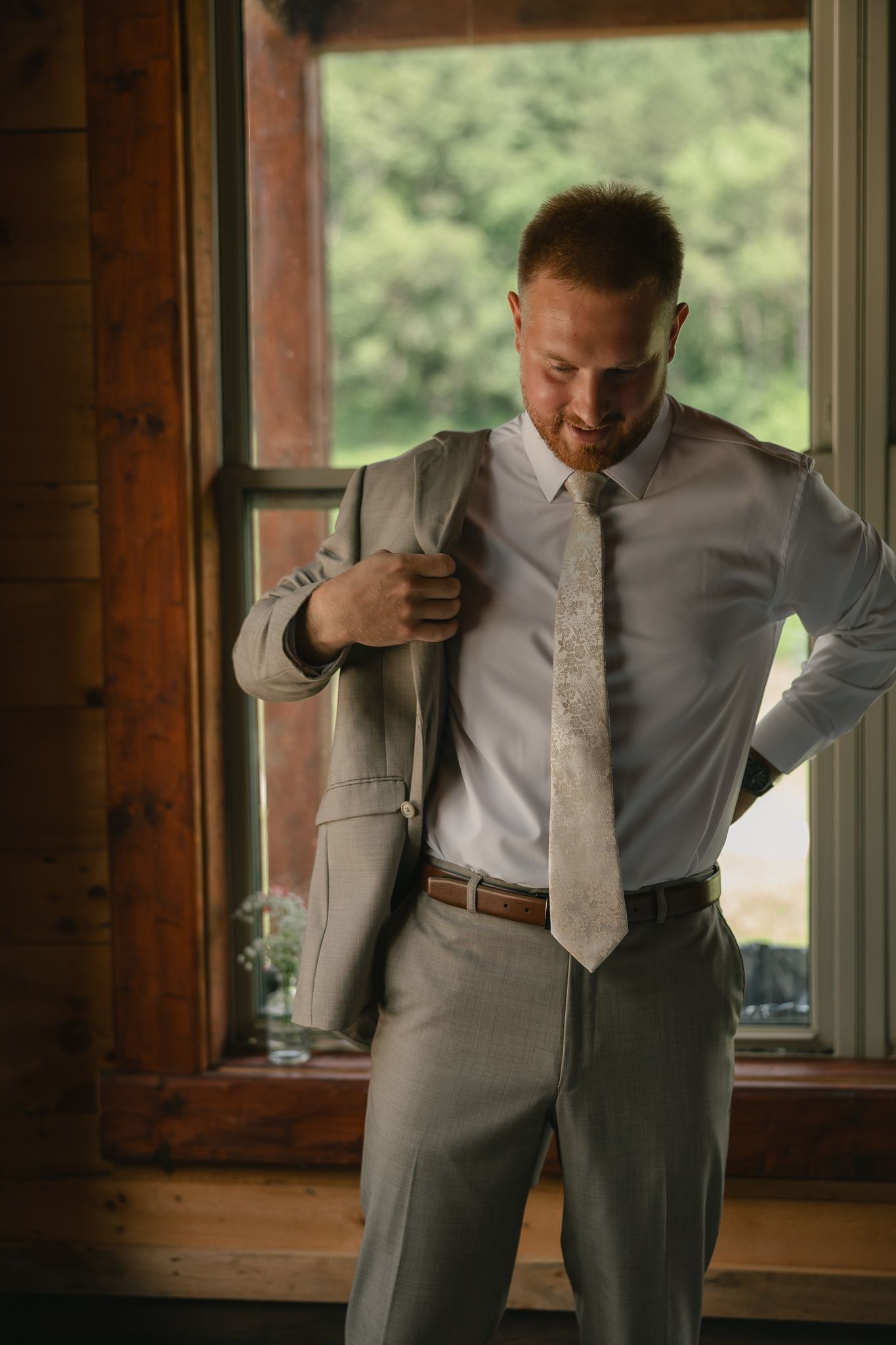 Groom adjusting formal attire by window in rustic cabin setting.