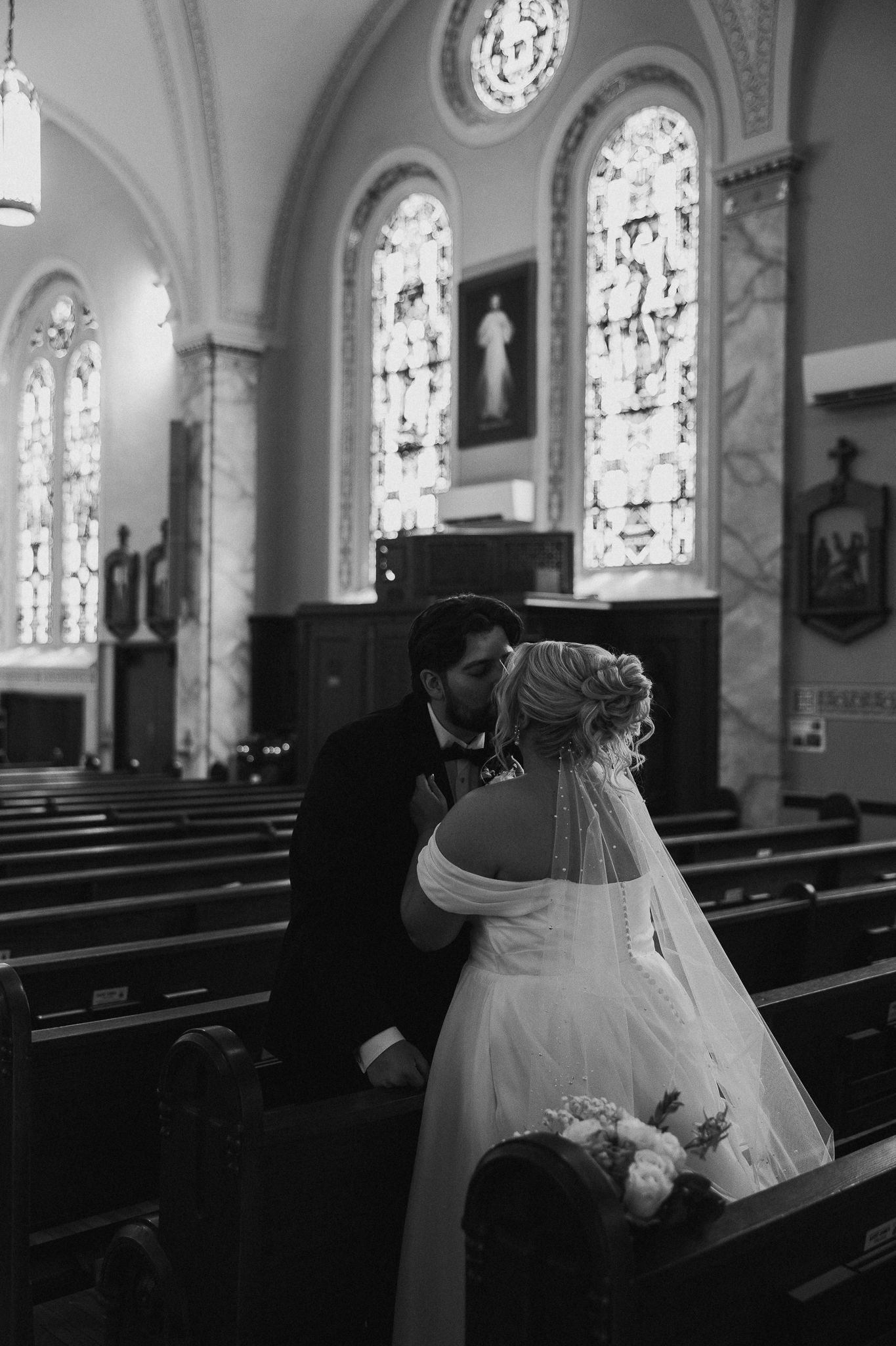 Black and white photo of a bride and groom sharing a kiss inside a beautiful church with stained glass windows.
