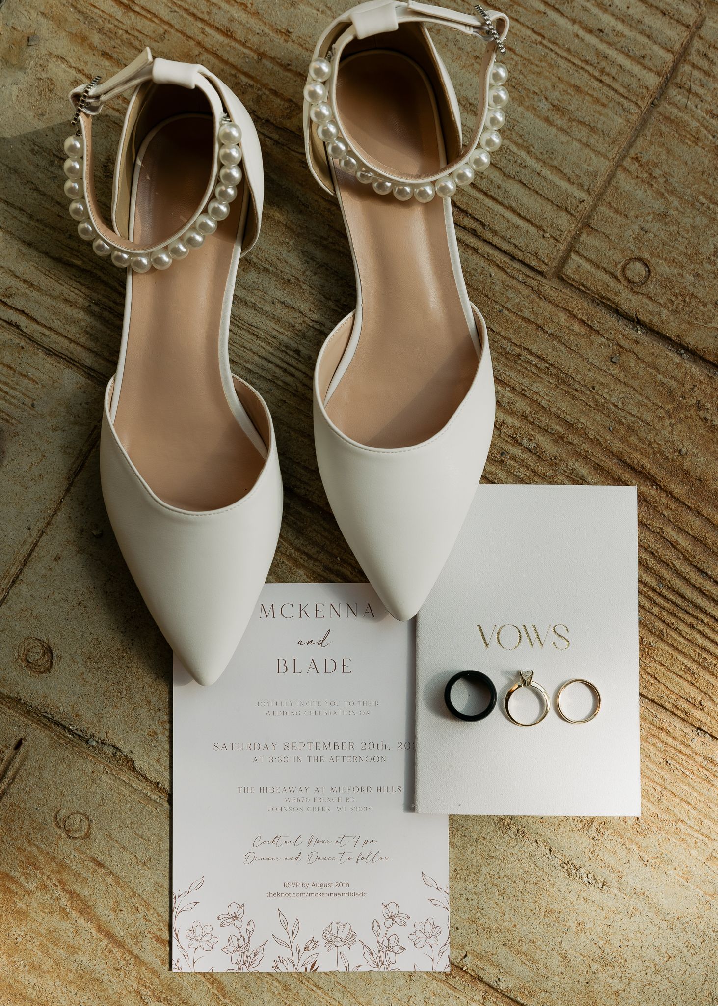 Wedding invitation suite with rings and white bridal shoes on rustic wooden surface.