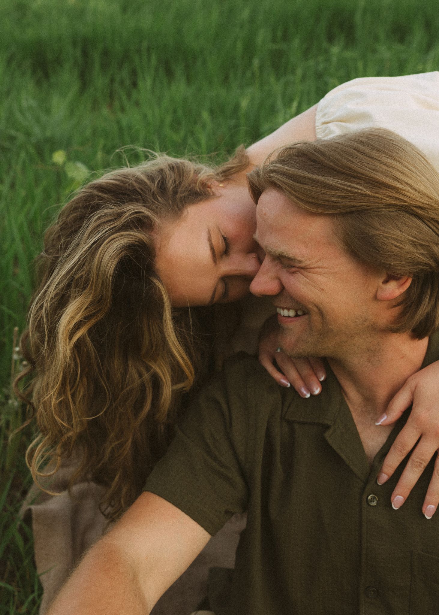 A couple laughs and nuzzles together in a lush green field during a romantic outdoor photo session.