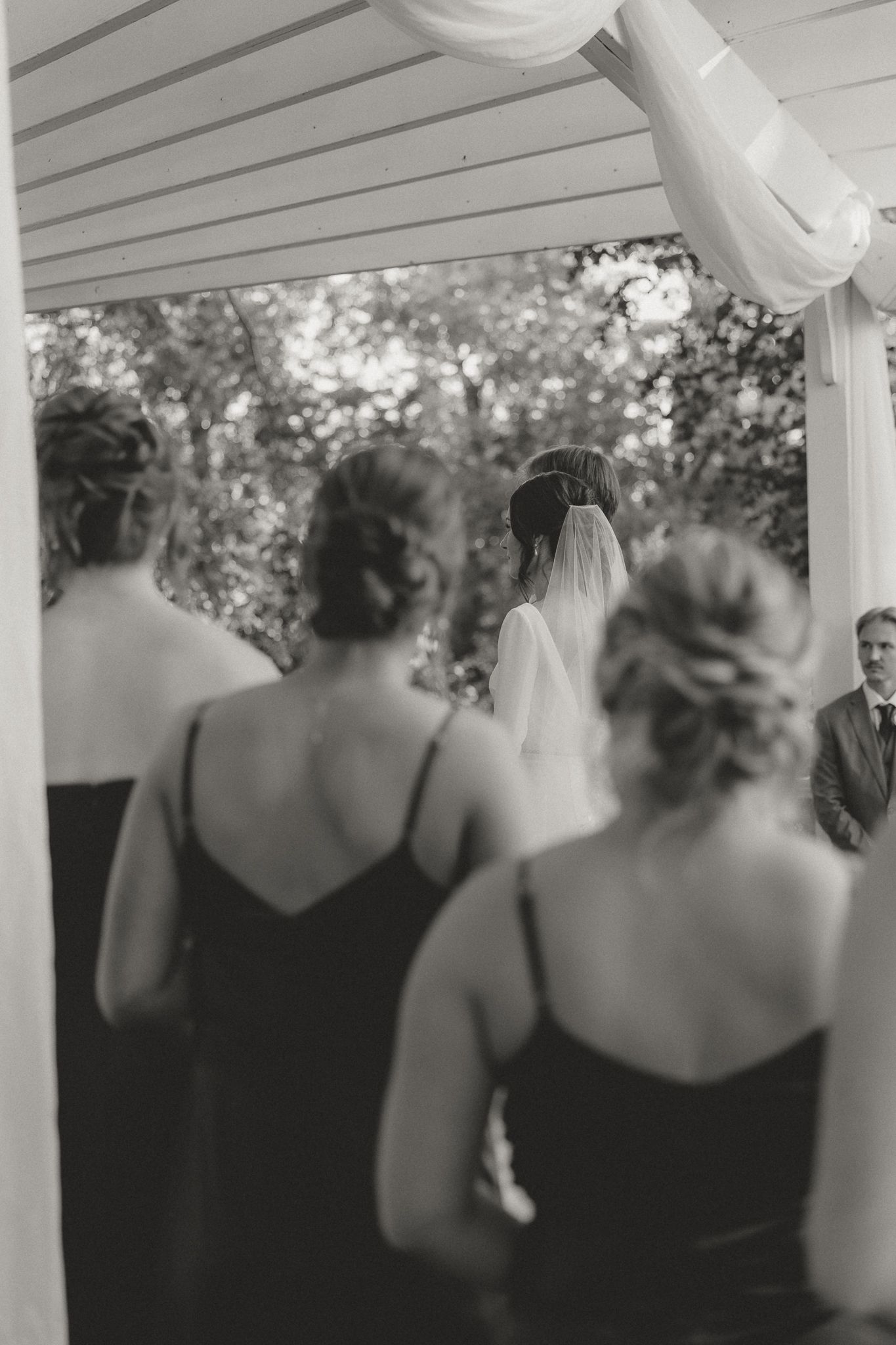 Bridesmaids in black dresses standing under white pergola at outdoor venue.