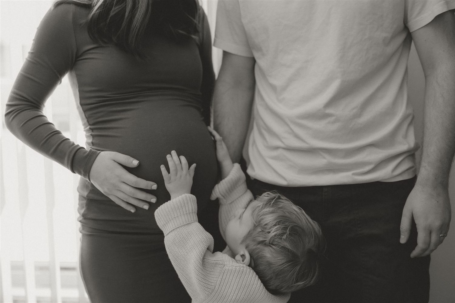 Small child touches pregnant mother's belly while standing with parents.