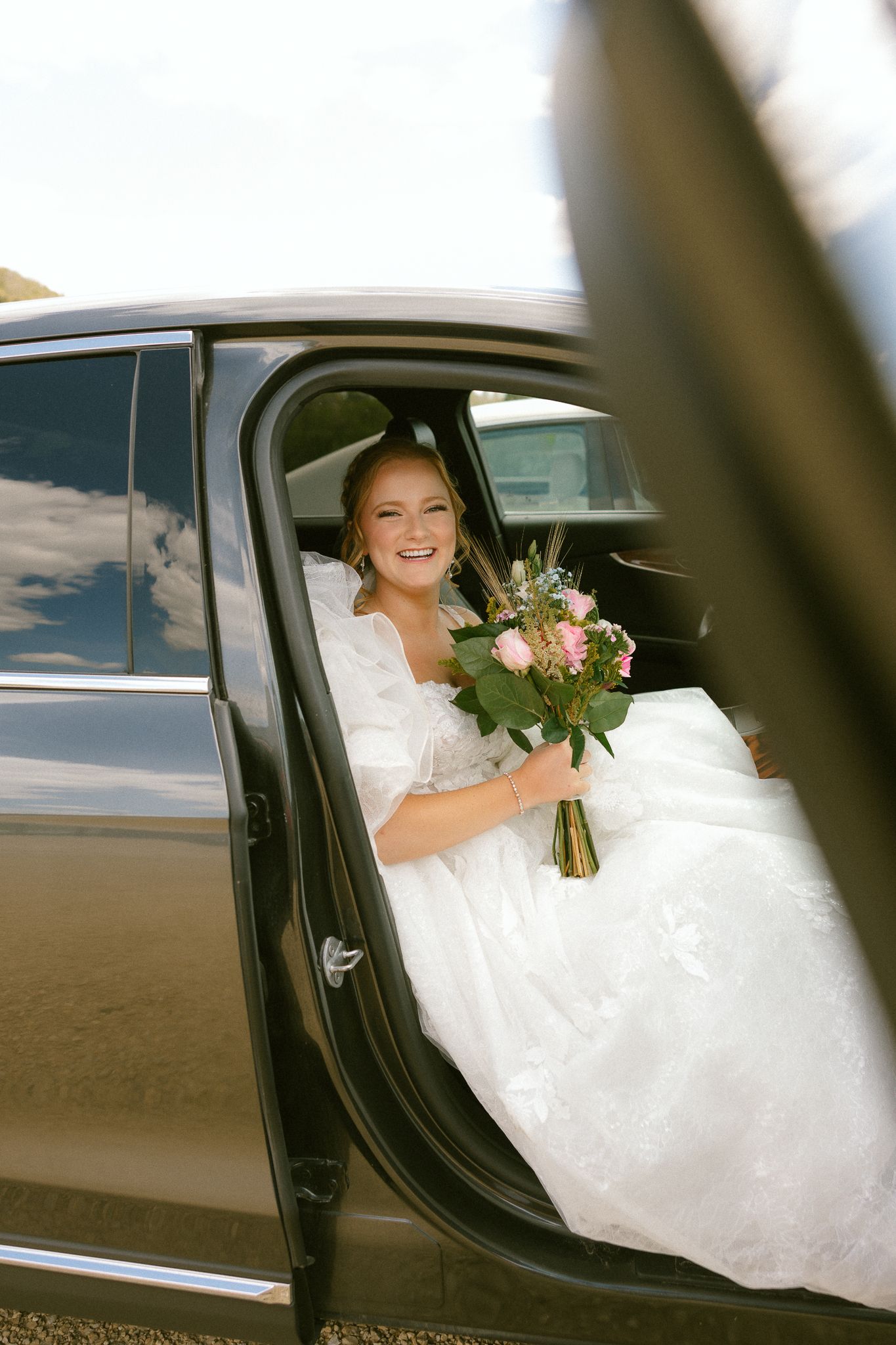 Smiling bride in white wedding dress holding pink flower bouquet sitting in black car with open door.