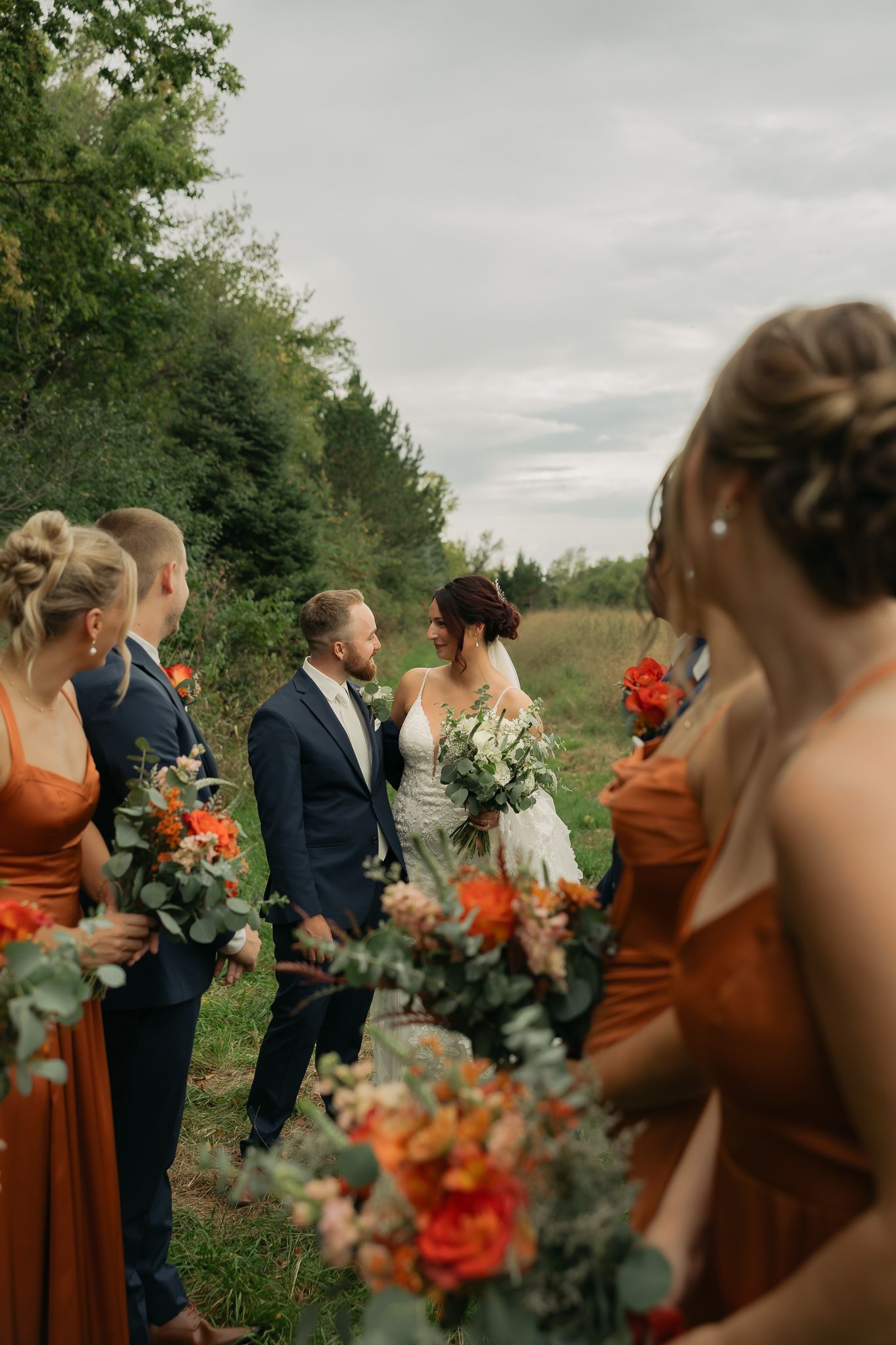 Wedding party holds bouquets of orange and white flowers while walking through a field.