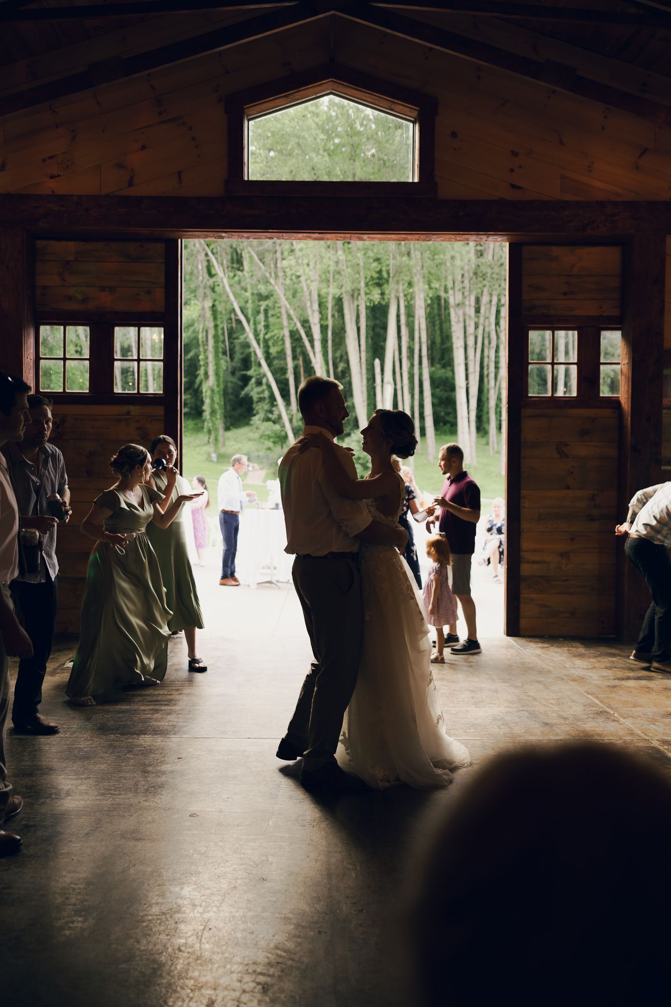 Silhouettes of wedding guests dancing in a rustic wooden barn with natural light streaming through windows.