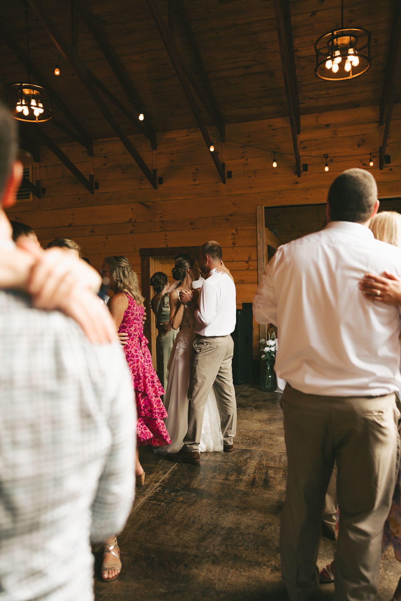 Wedding guests dance and celebrate on a rustic wooden dance floor under warm lighting at a reception.