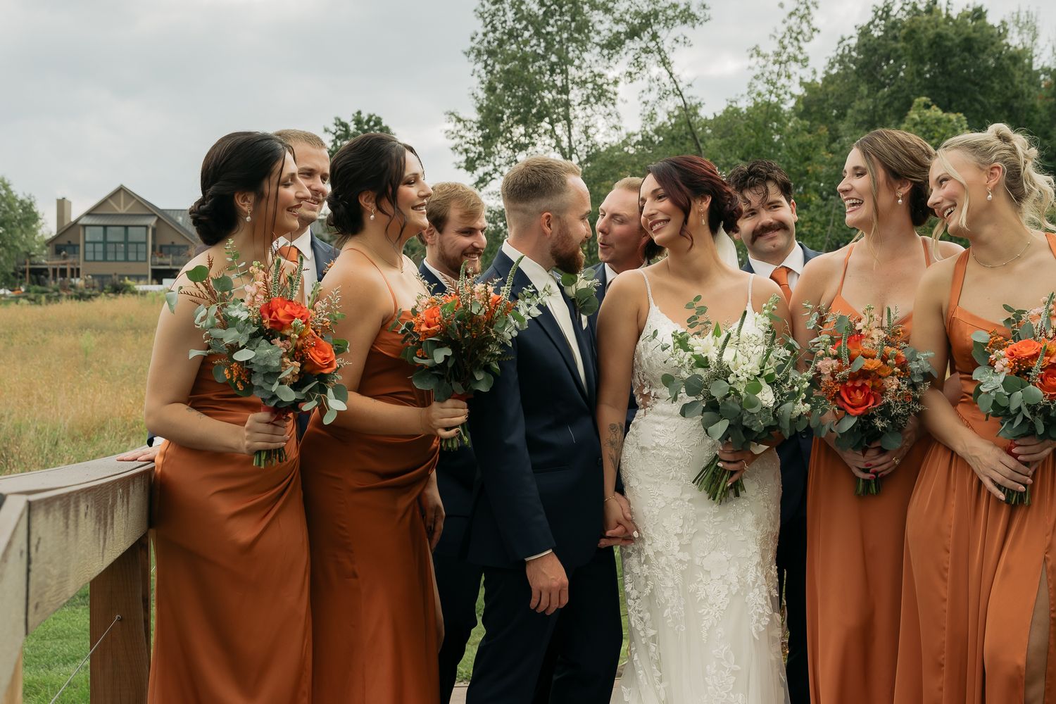 Wedding party in rust-colored dresses and dark suits poses outdoors.