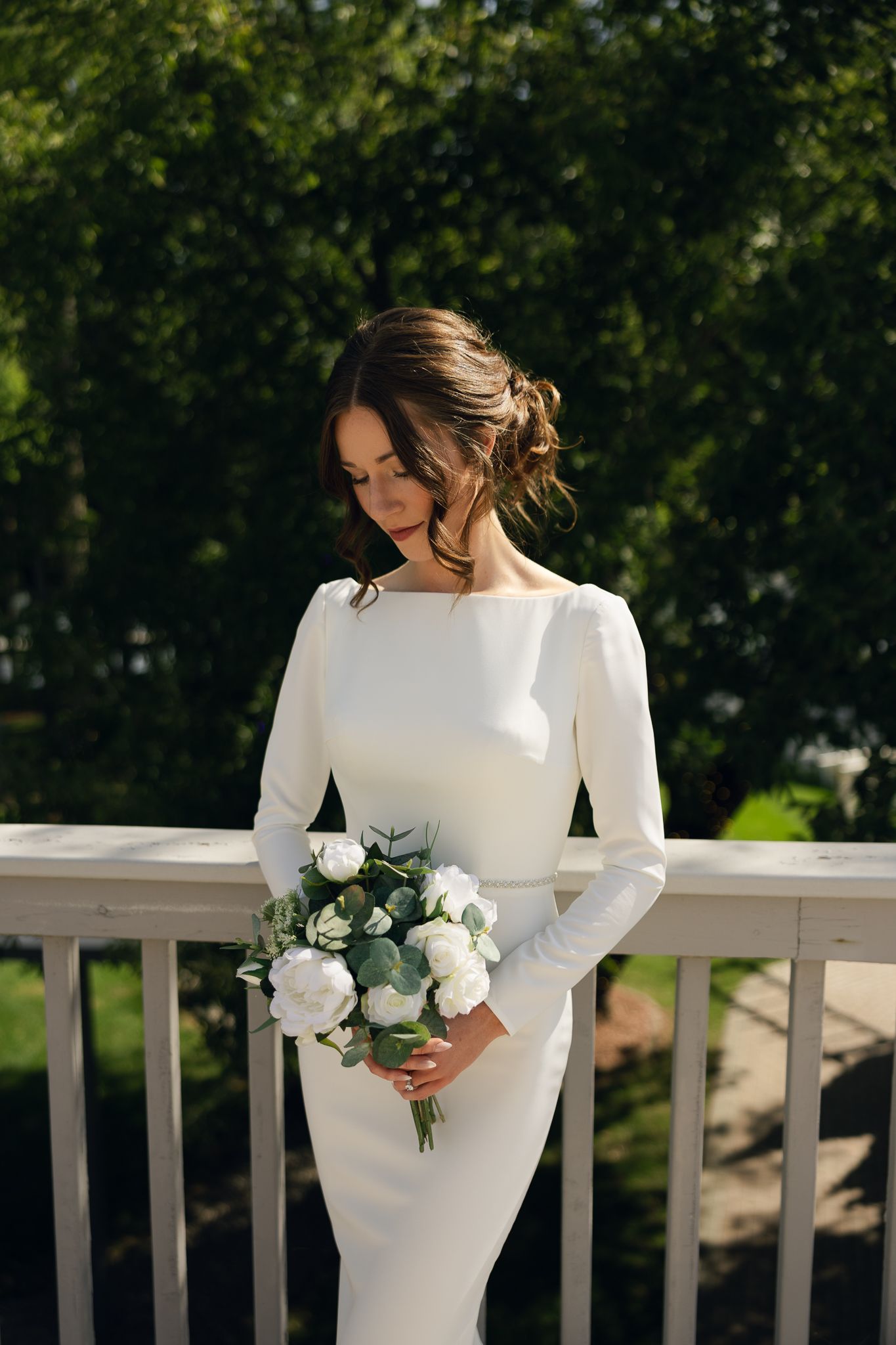 Bride in sleek white dress holds classic white and green bouquet on sunny balcony.
