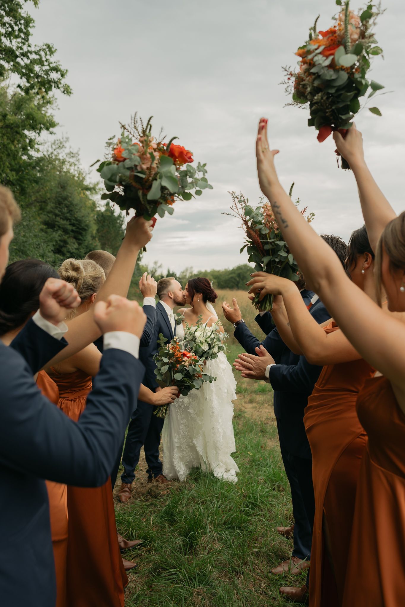Wedding party raises bouquets in celebration during outdoor ceremony with bridesmaids in rust-colored dresses.